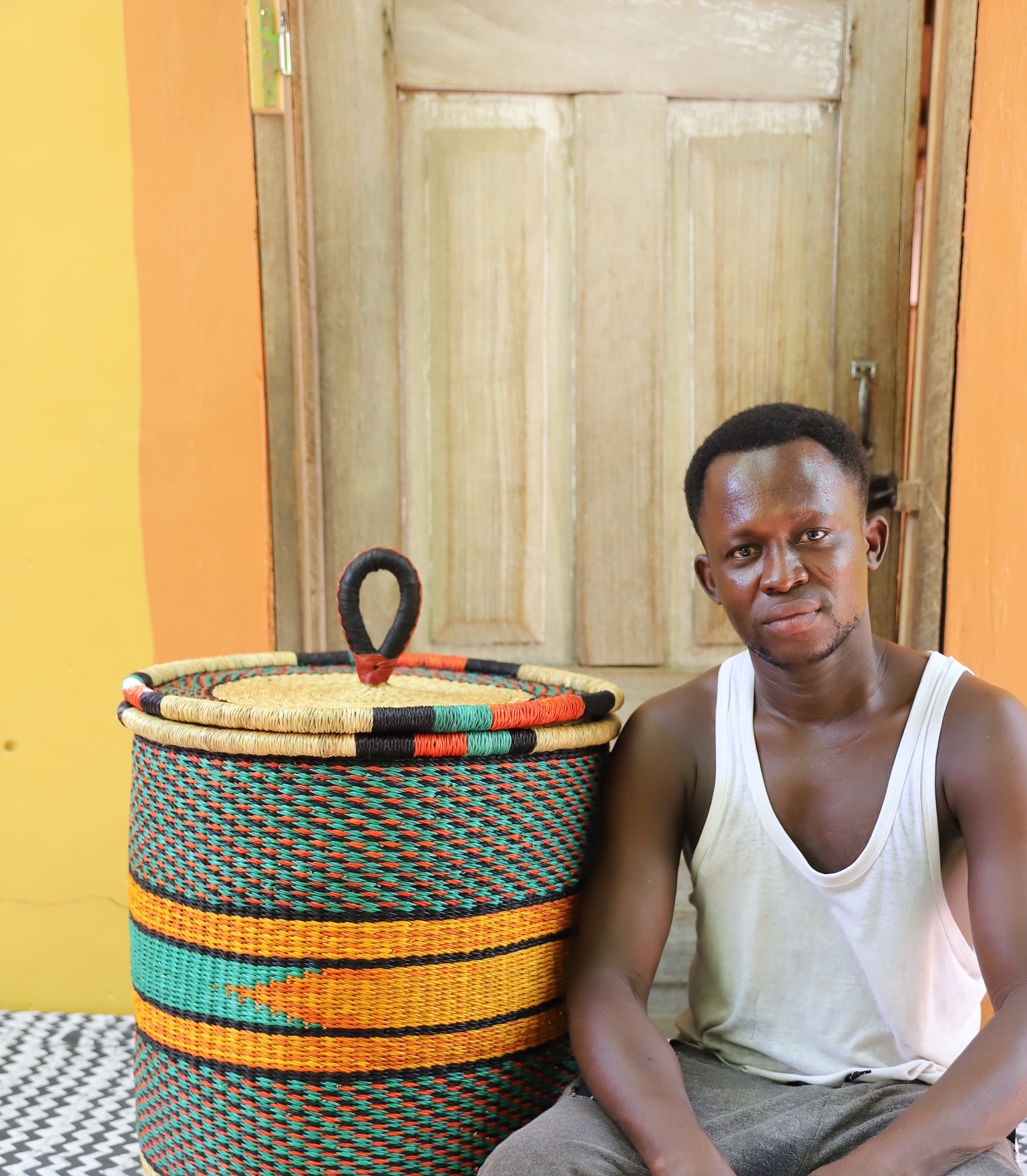 Large coiled-rim lidded laundry basket in dark weave with orange and black banding, shown beside a seated person for scale.