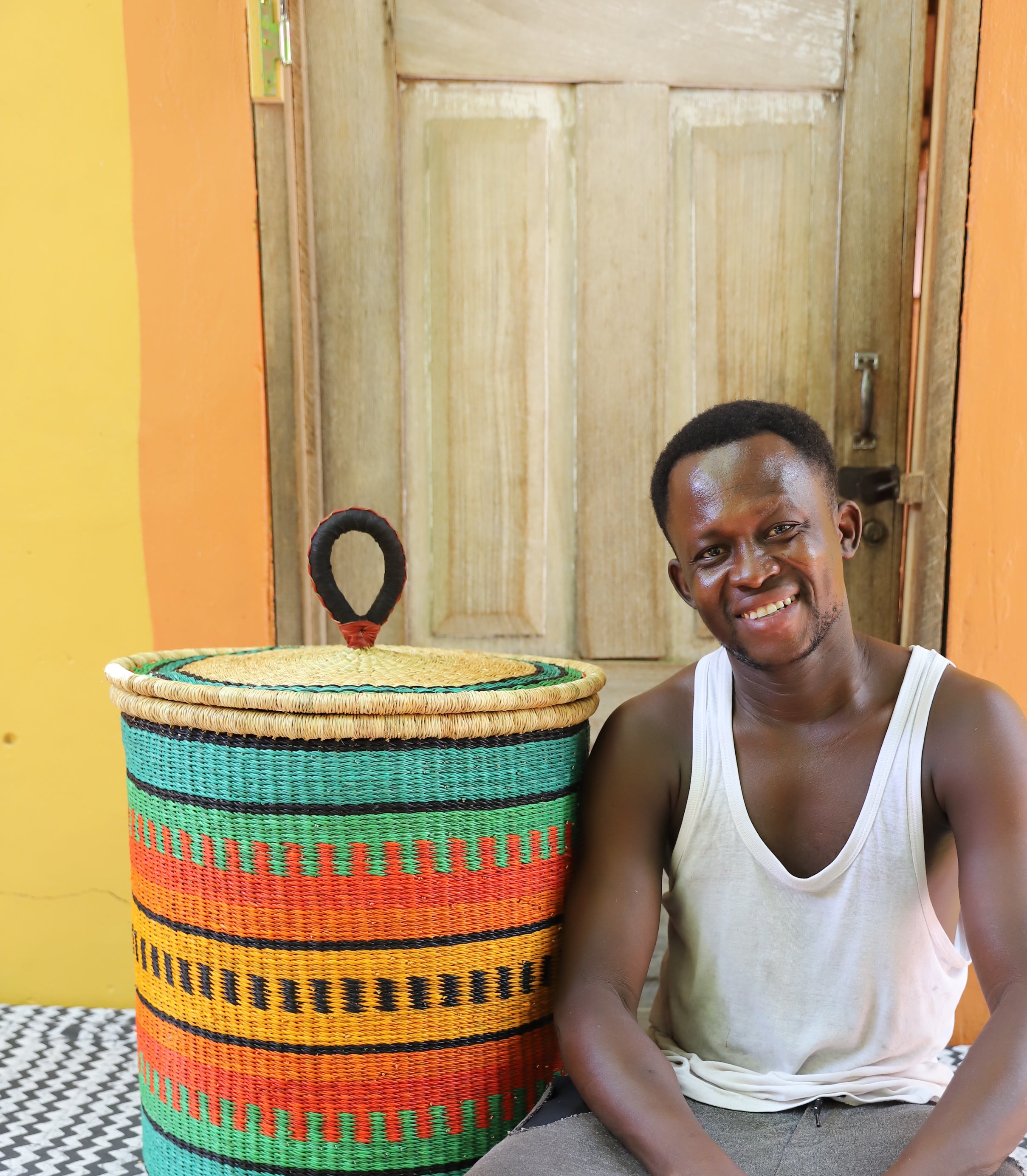 Large coiled-rim lidded laundry basket in green, orange and black striped weave, shown beside a seated person for scale.