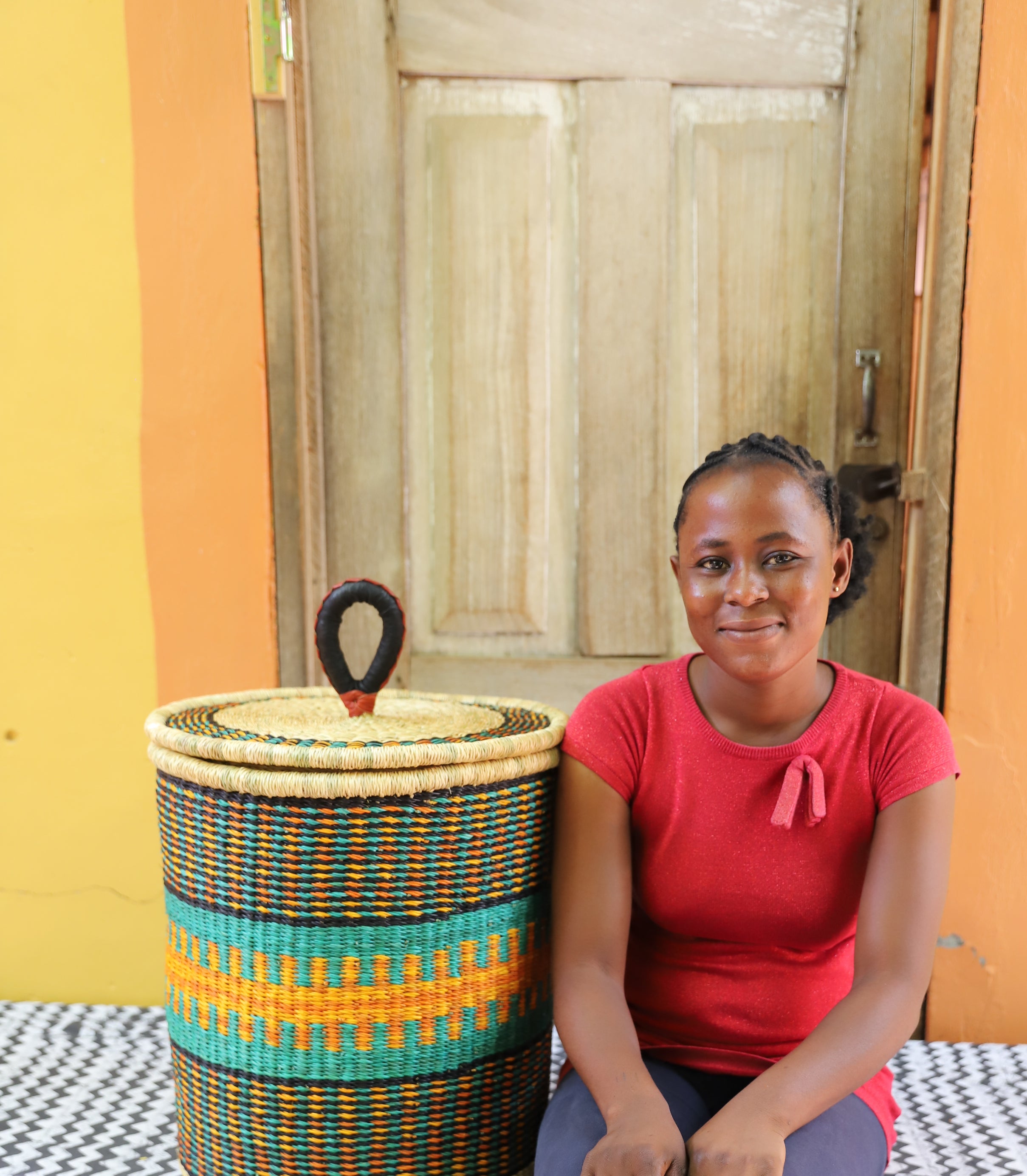 Coiled-rim lidded laundry basket in dark weave with turquoise and orange banding, shown beside a seated person for scale.