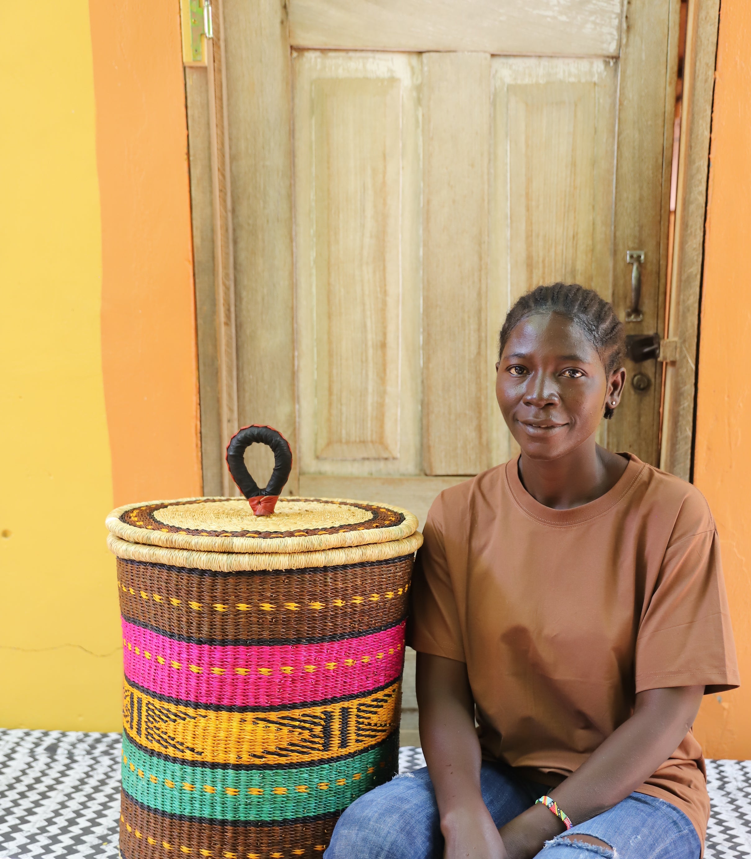 Coiled-rim lidded laundry basket in brown, pink, mustard and teal striped weave, shown beside a seated person for scale.