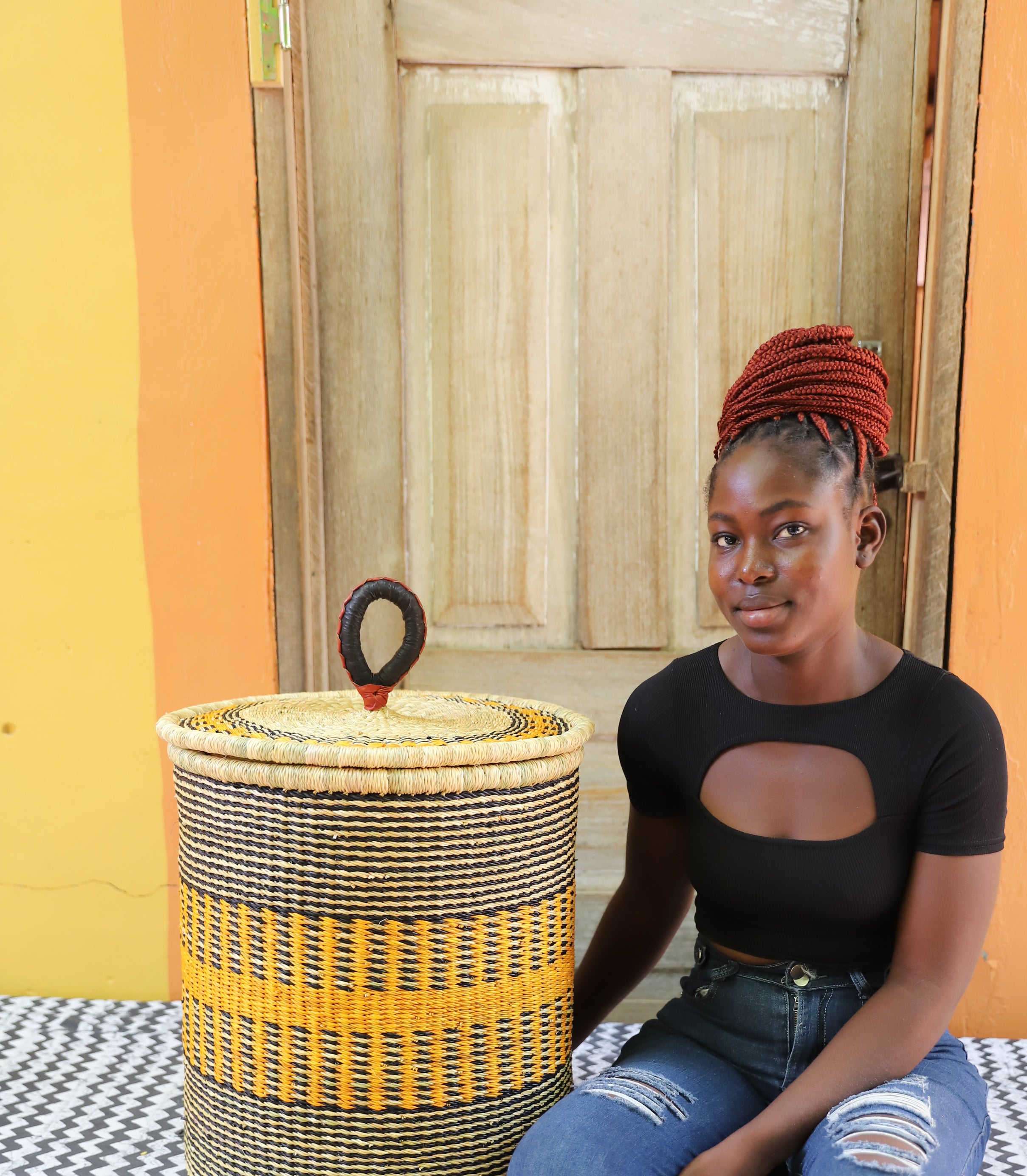 Coiled-rim lidded laundry basket in natural straw with a striped orange band, shown beside a seated person for scale.