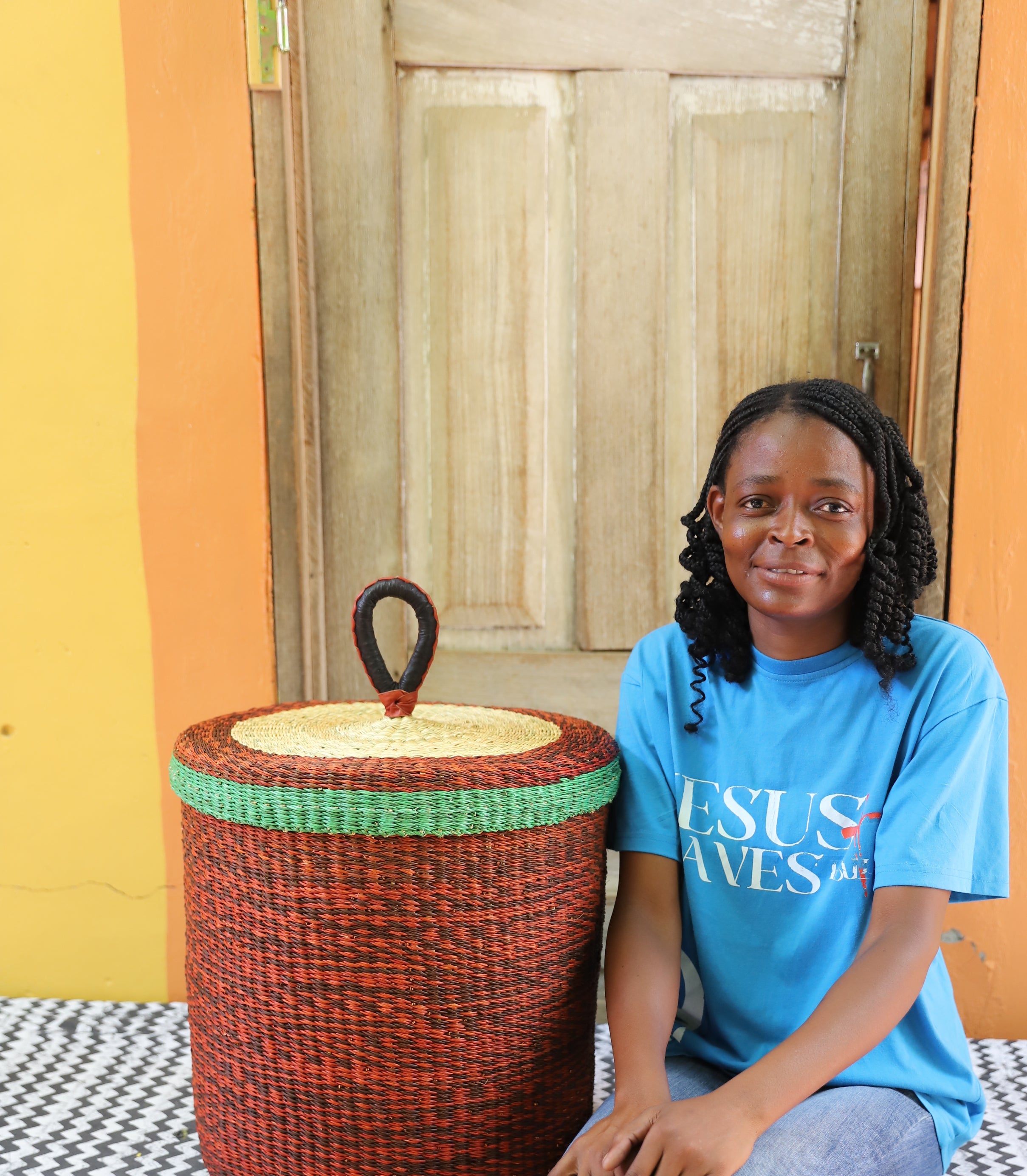 Lidded laundry basket in solid rust-red weave, shown beside a seated person for scale.