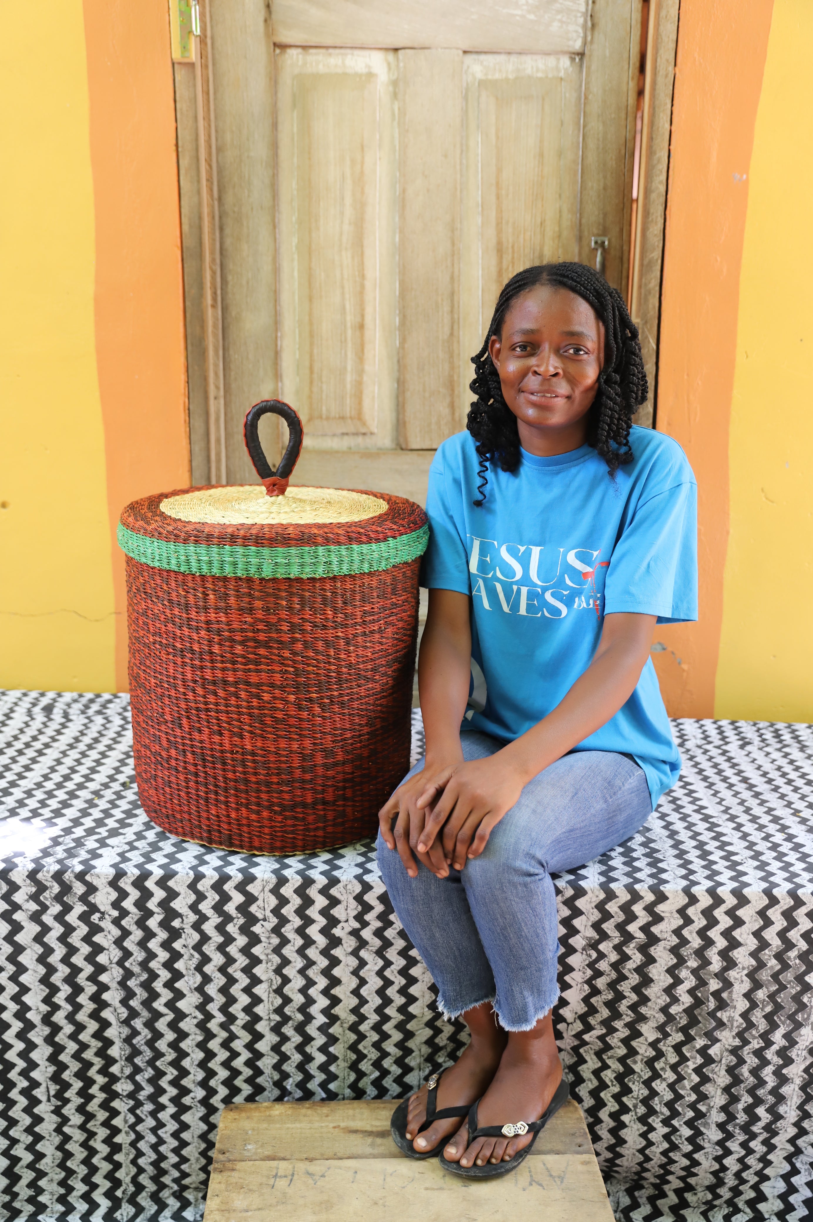 Lidded laundry basket in solid rust-red weave, shown beside a seated person for scale.