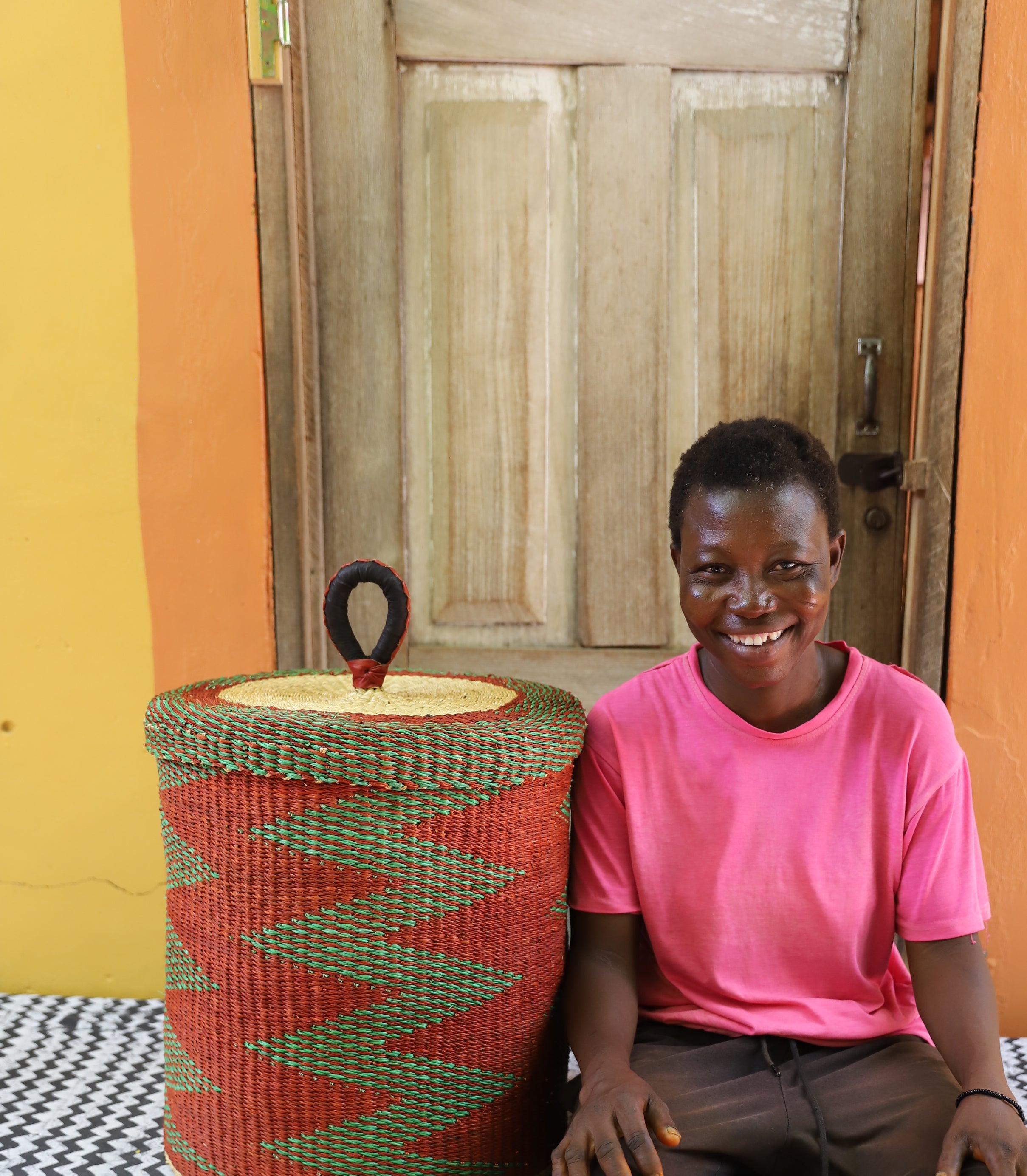 Lidded laundry basket in rust and green zigzag weave, shown beside a seated person for scale.
