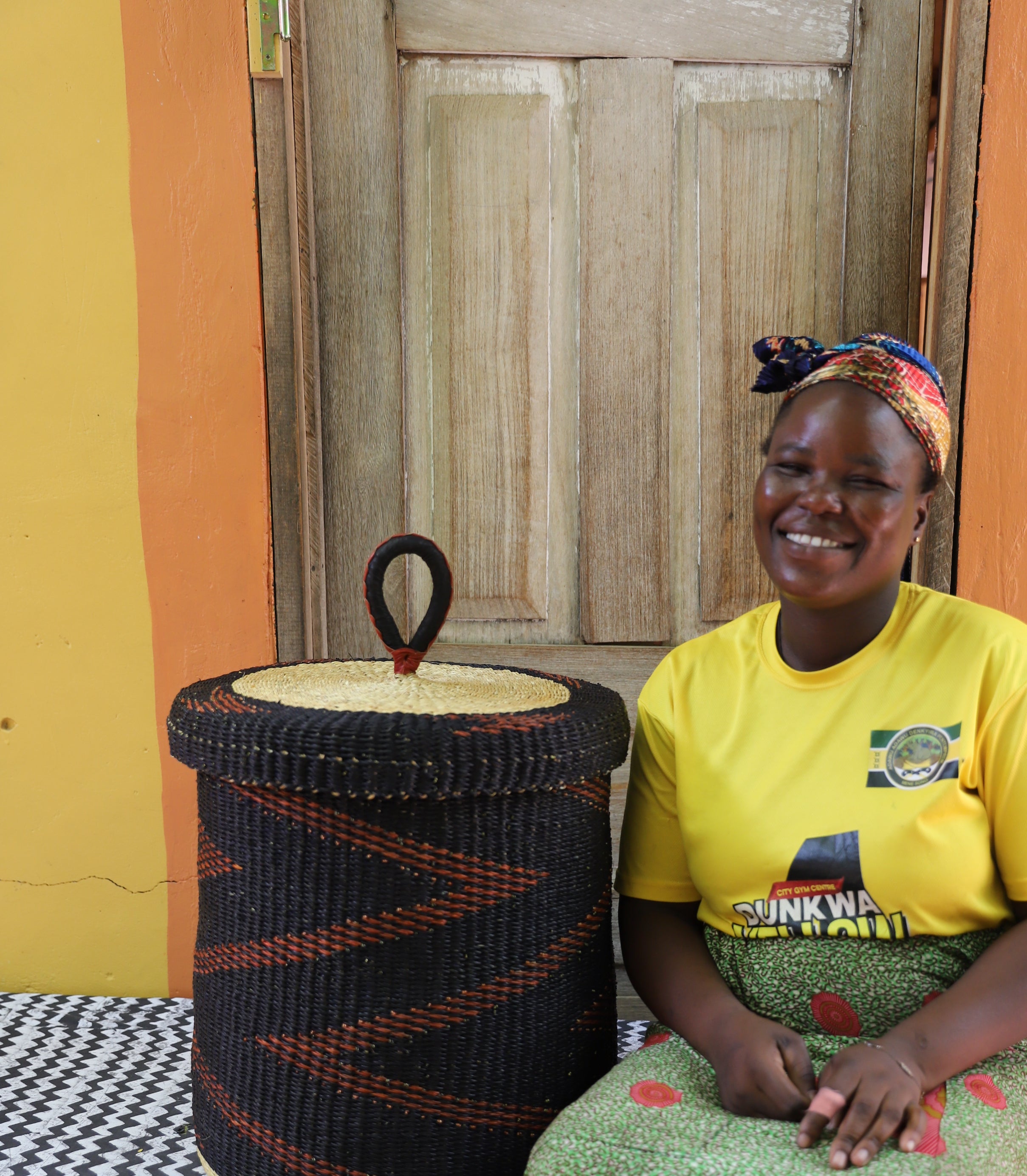 Lidded laundry basket in dark brown elephant grass, shown beside a seated person for scale.