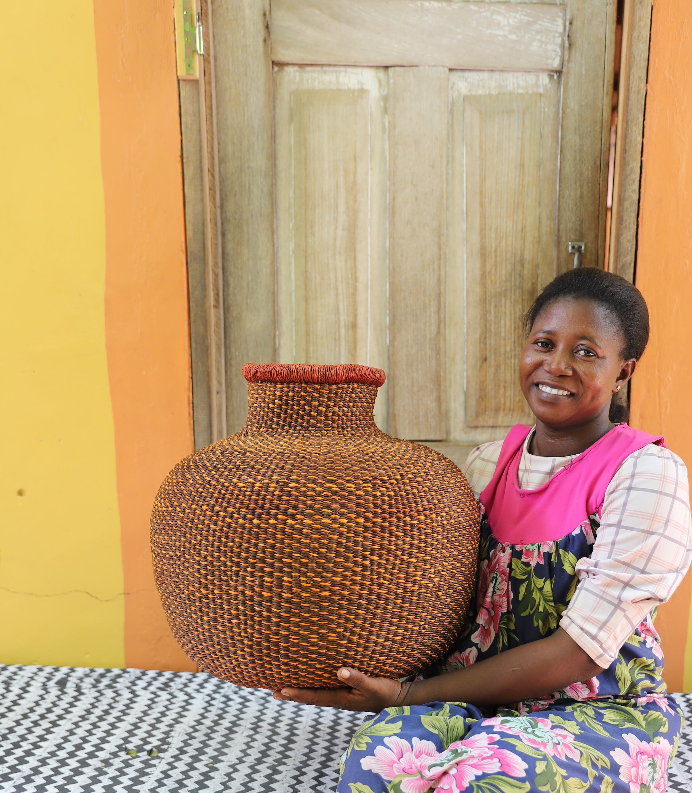 Bulbous décor basket in natural straw with a rust-dyed neck band, held by a seated person for scale.