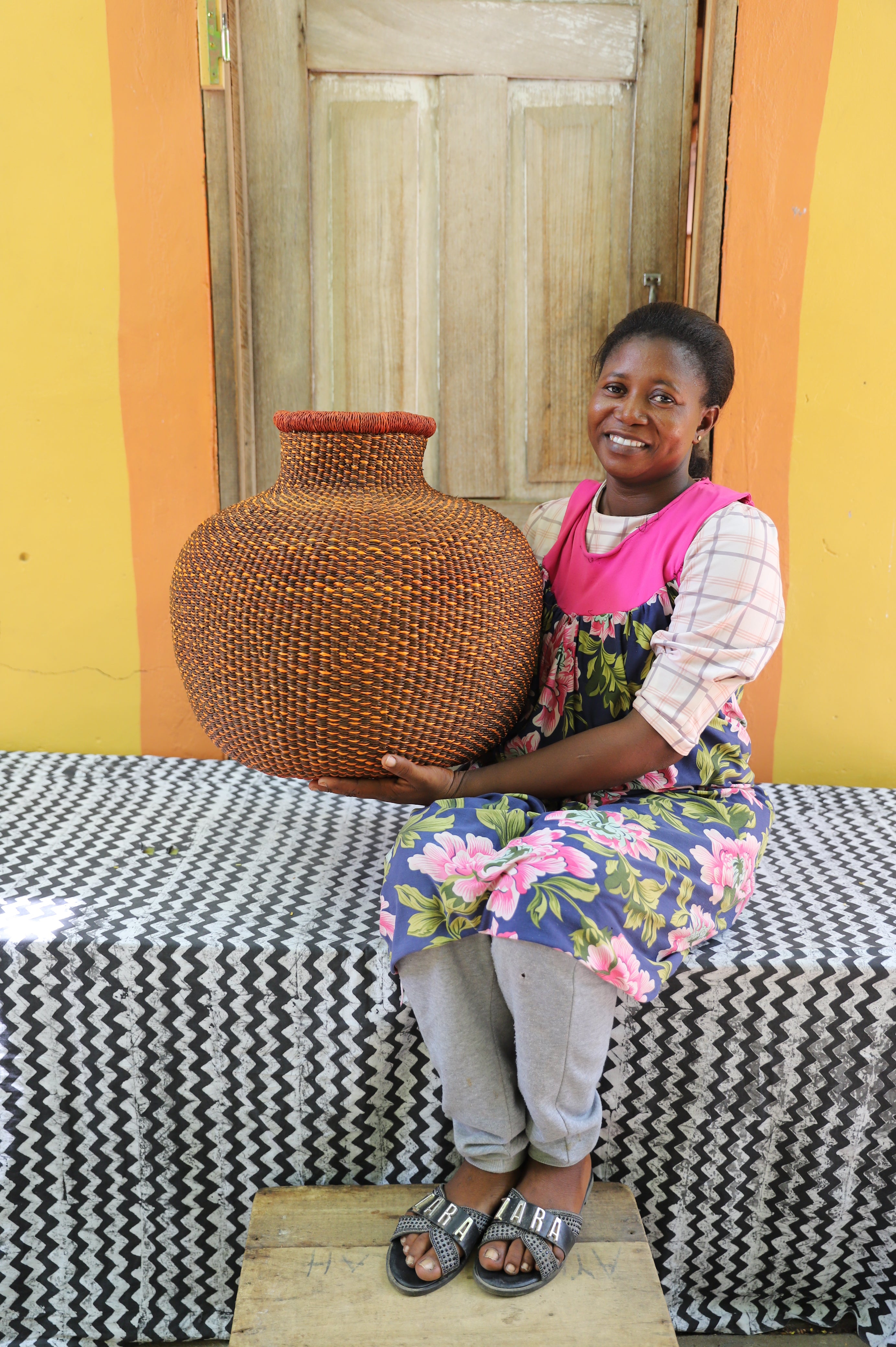 Bulbous décor basket in natural straw with a rust-dyed neck band, held by a seated person for scale.