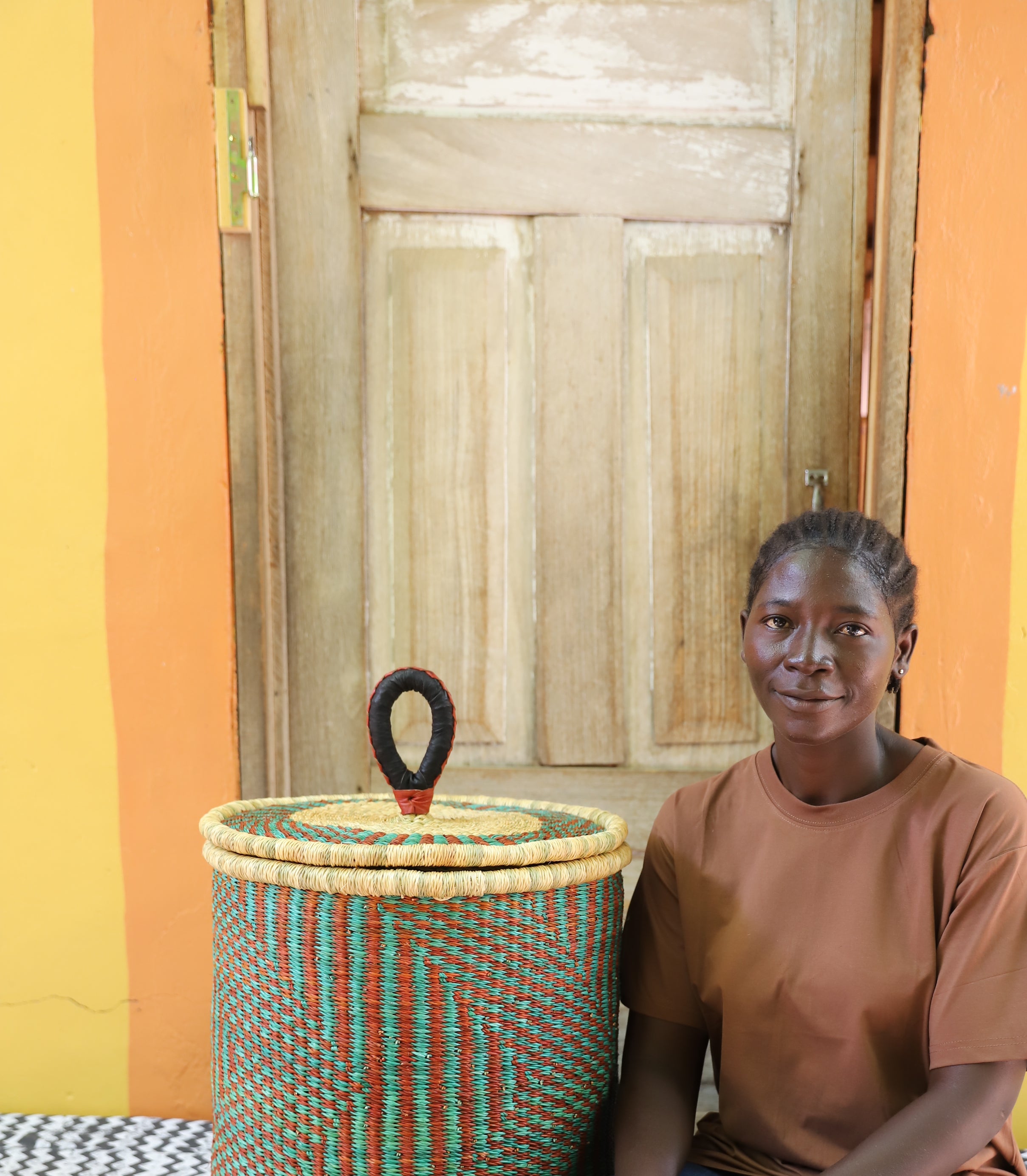Coiled-rim lidded laundry basket in natural straw with teal and maroon banding, shown beside a seated person for scale.