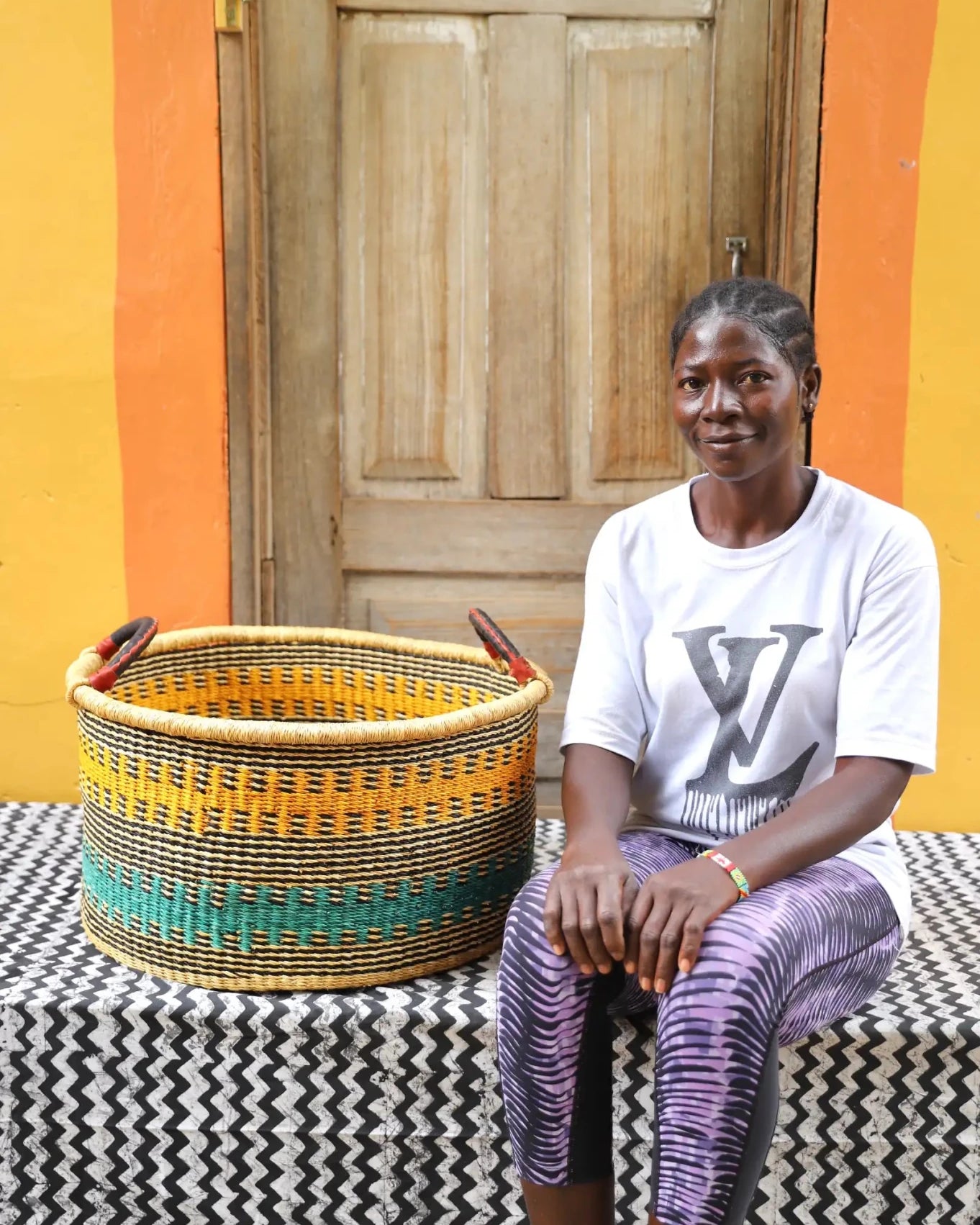 Woven Tub (Large) with woman weaver handwoven in Ghana sitting on black and white cloth and wooden door in background