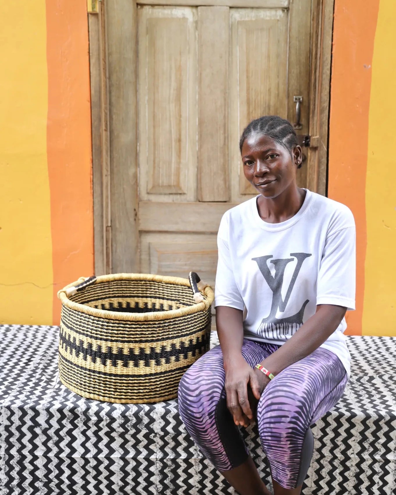 Woven Tub (Small) with woman weaver handwoven in Ghana sitting on black and white cloth and wooden door in background