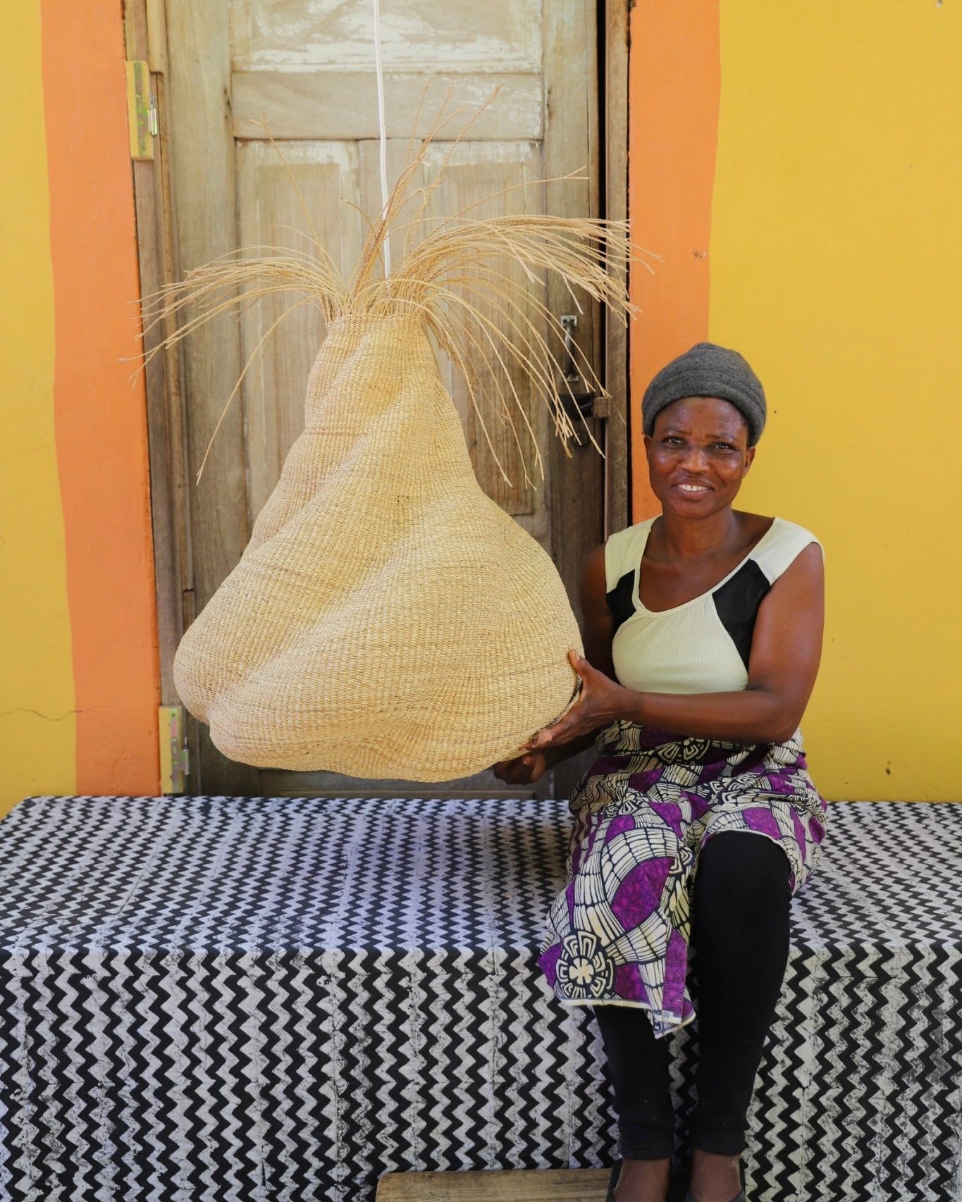 Woman sitting on a bench with a woven basket in front of a yellow wall.