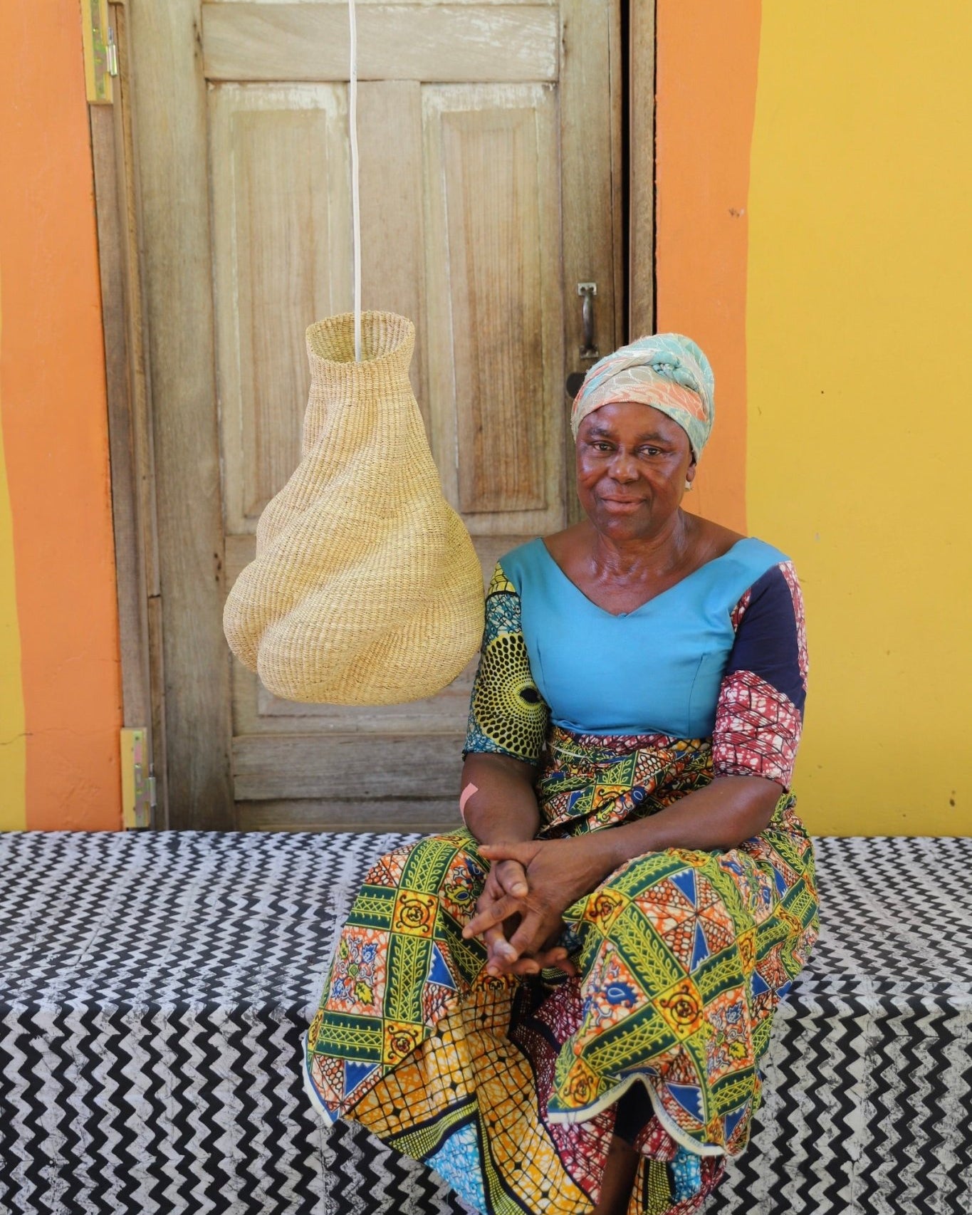 Woman sitting on a bench in front of a colorful wall with a woven lamp hanging.