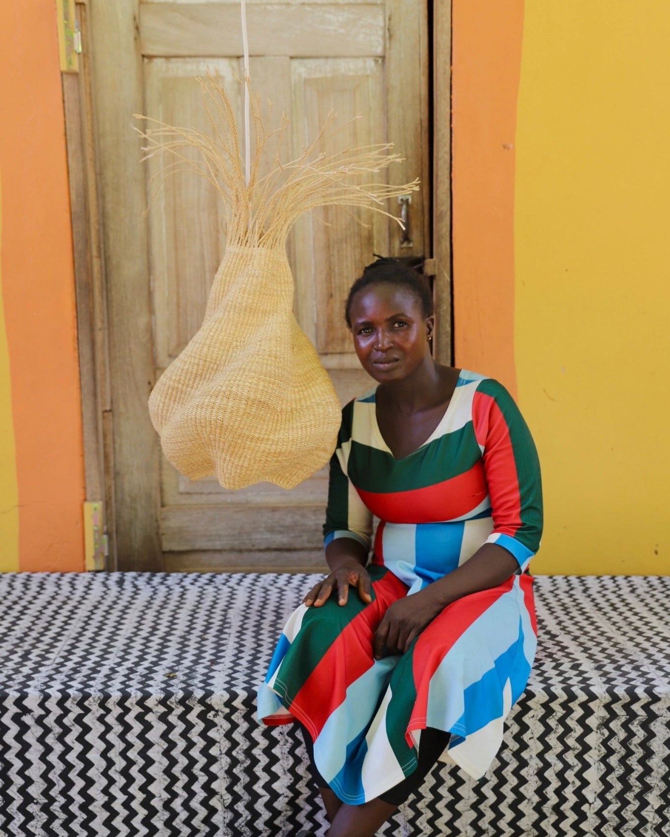 Woman sitting on a bed with a colorful dress in a room with a yellow wall and wooden door.