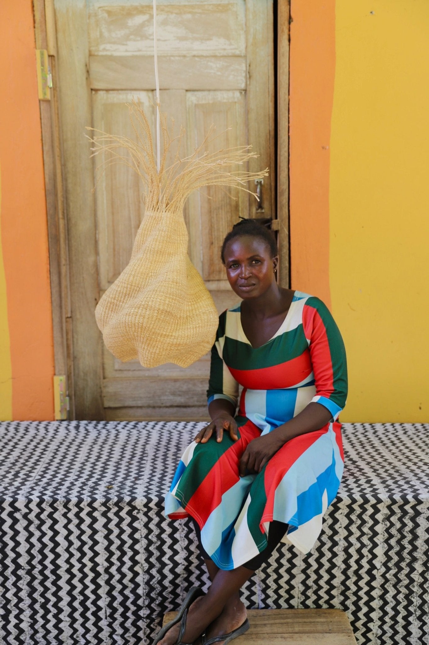 Woman sitting on a bed with a colorful dress in a room with a yellow wall and wooden door.