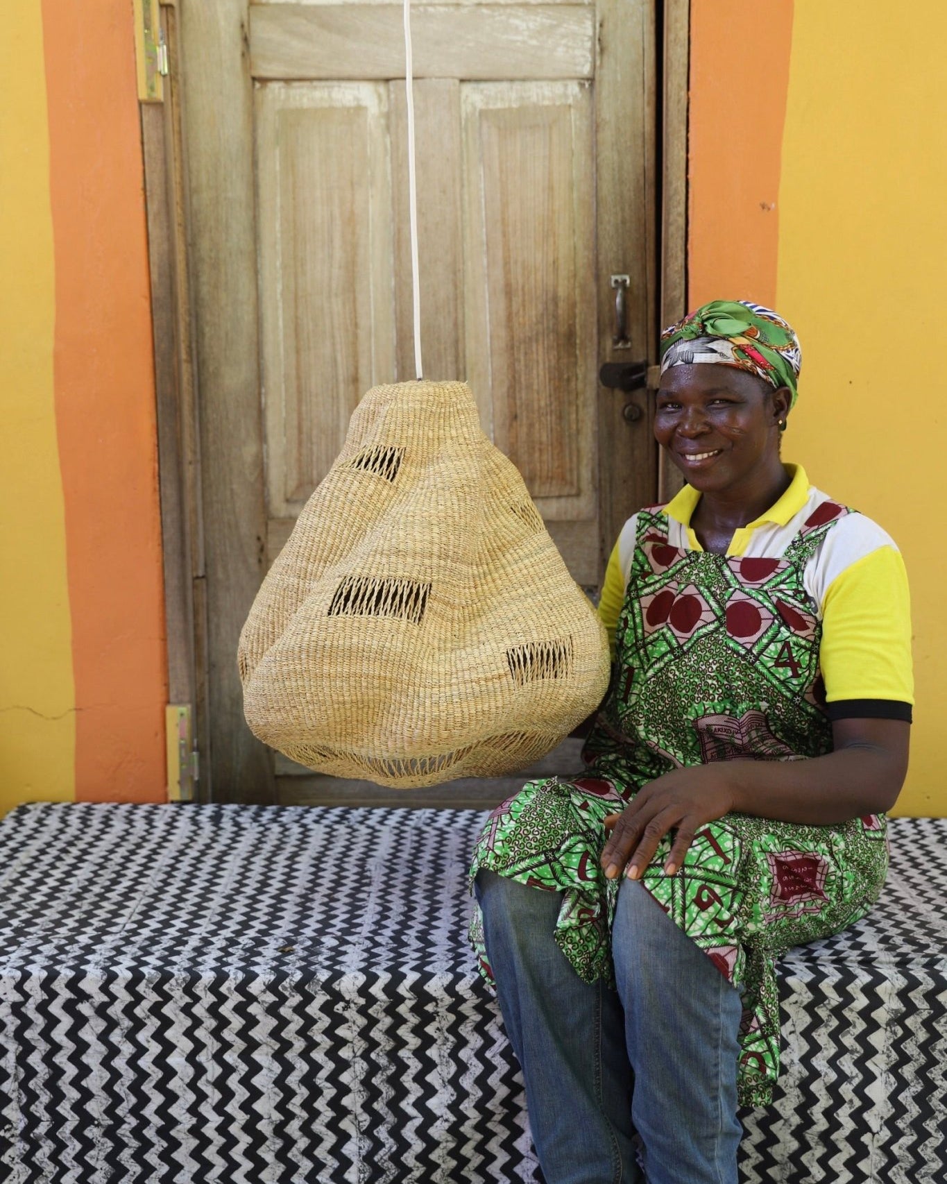 Woman sitting on a bench holding a woven basket against a colorful wall.