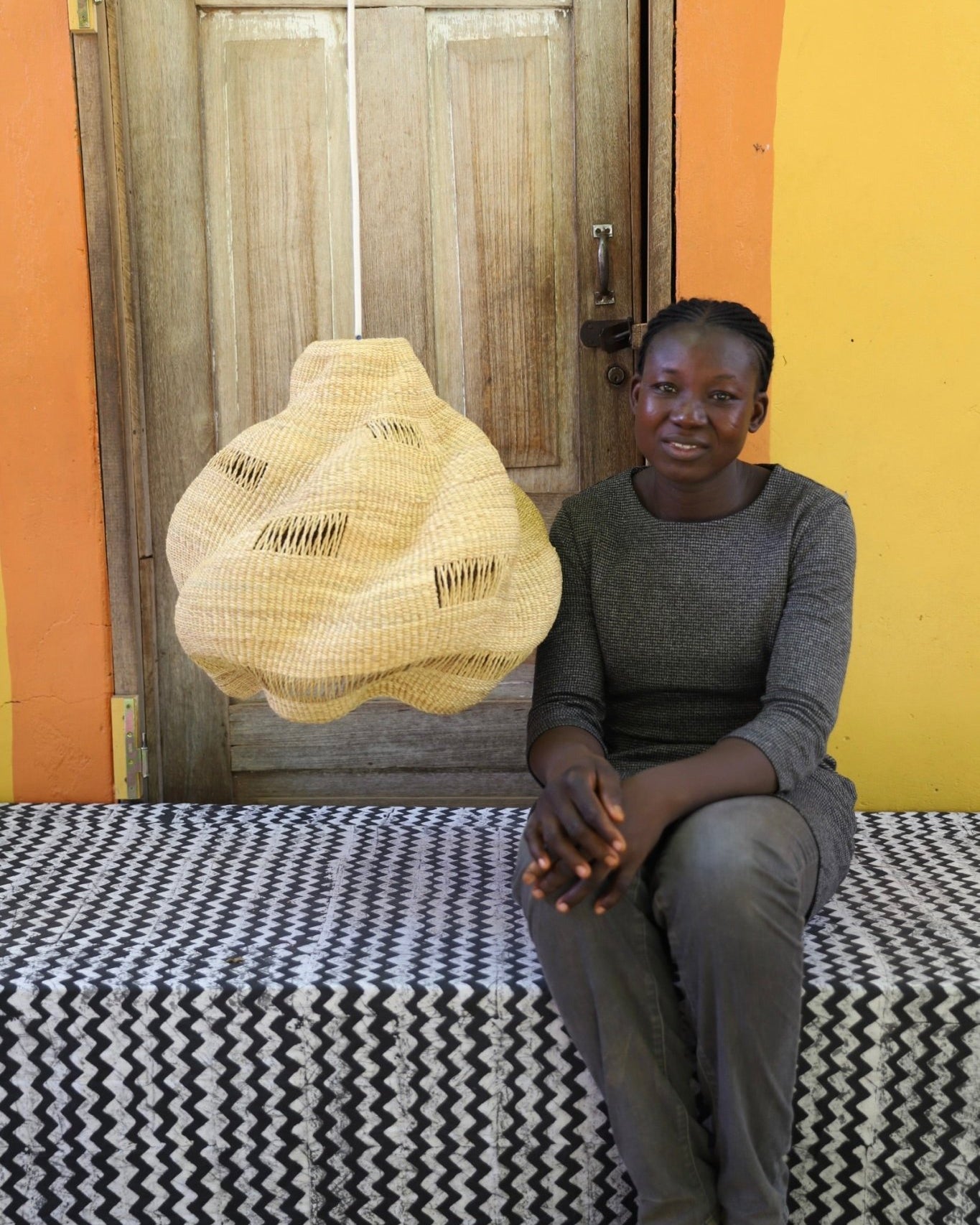 Woman sitting on a bed with a woven basket in front of a wooden door.