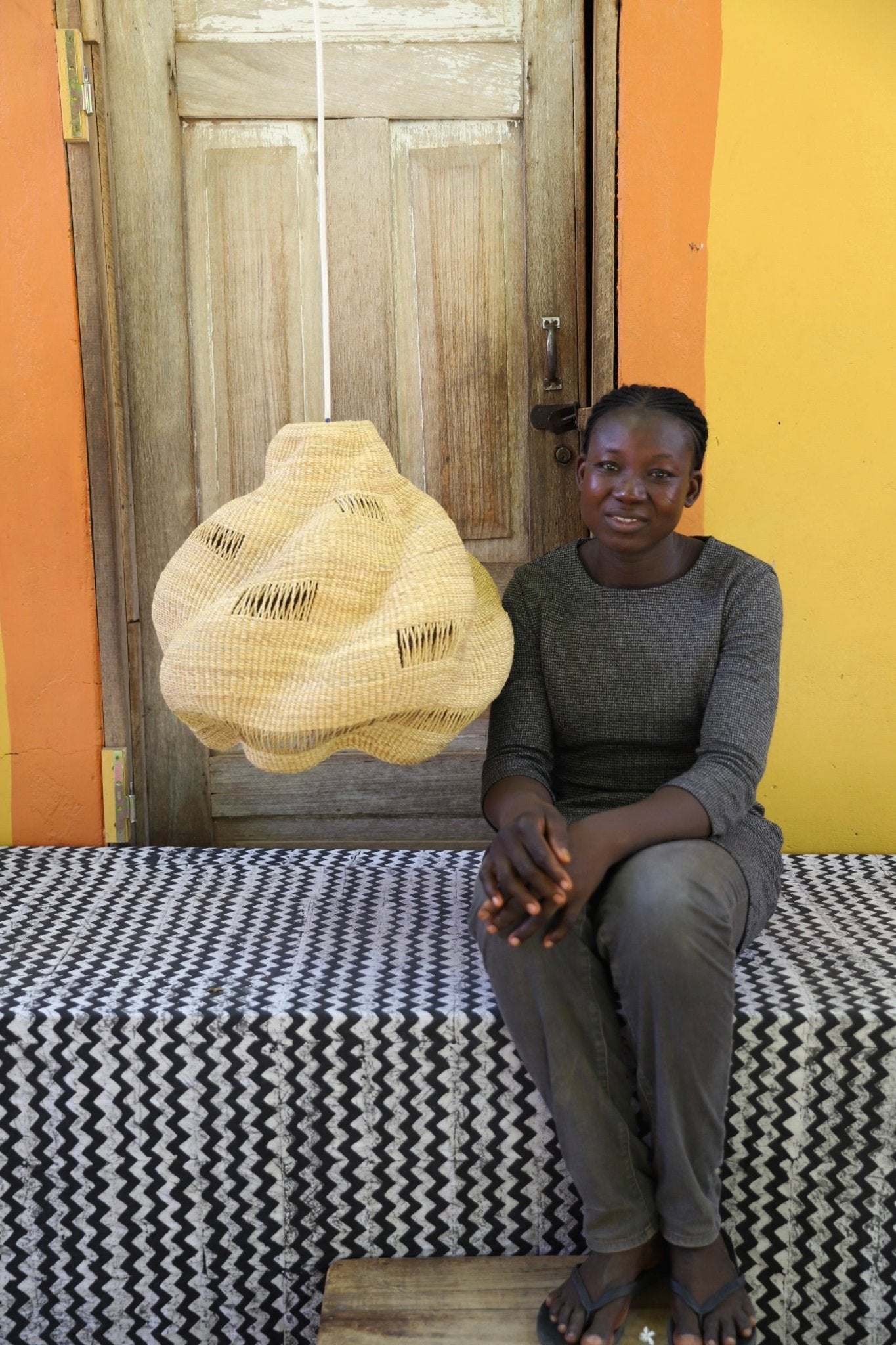 Woman sitting on a bed with a woven basket in front of a wooden door.