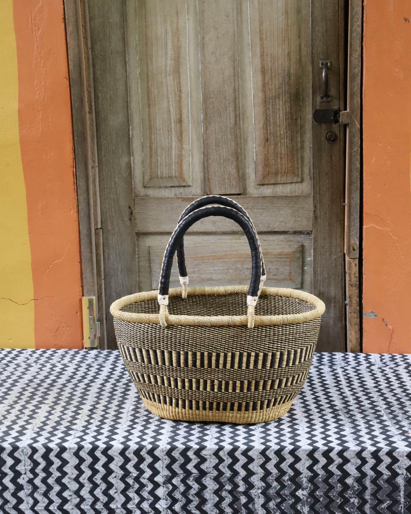 Woven carry on bag with premium leather handles on a black and white cloth against a wooden door.