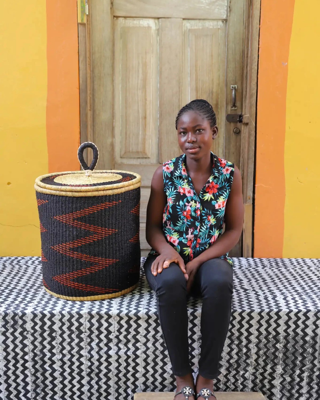 Woman sitting on a bench with a woven basket next to her against a colorful wall.