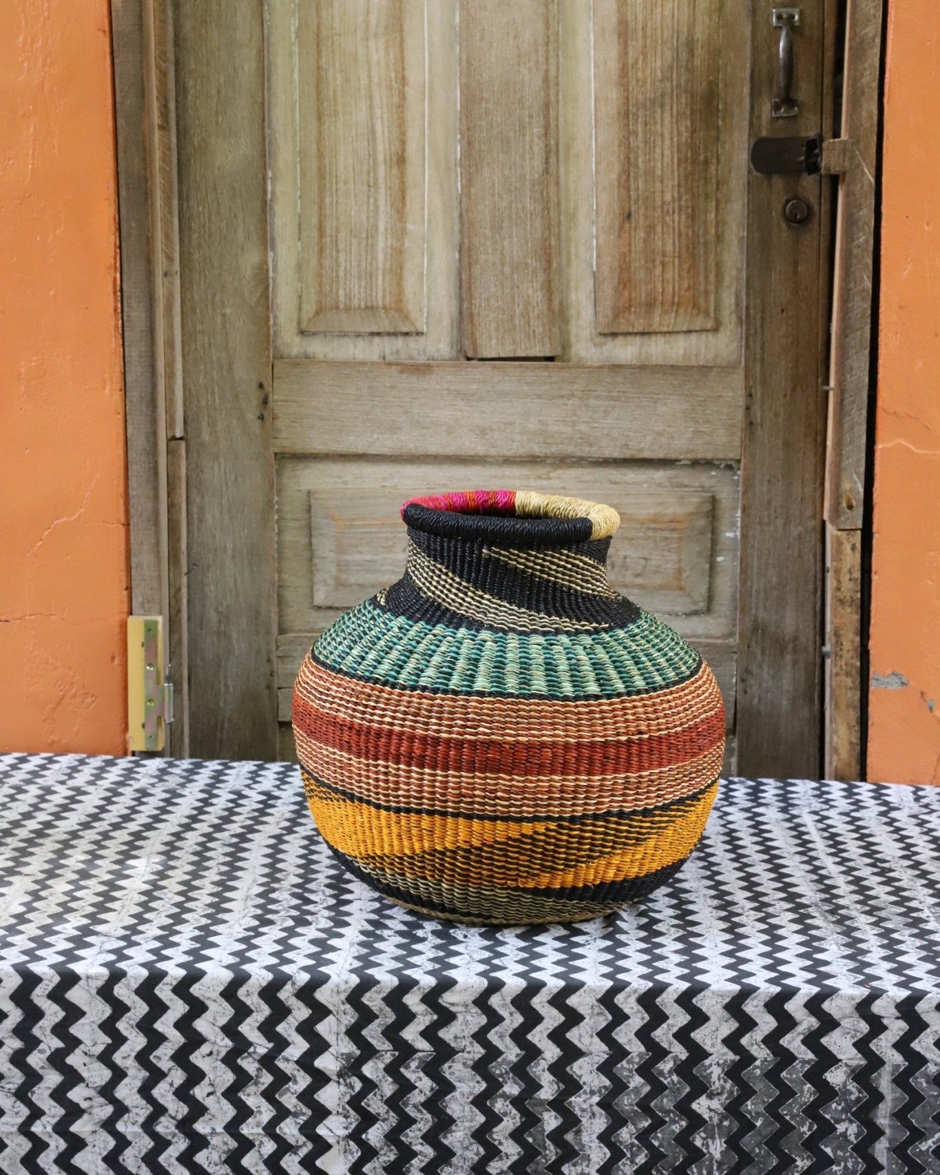 Decorative handwoven basket with colorful patterns on a black and white cloth against a wooden door.