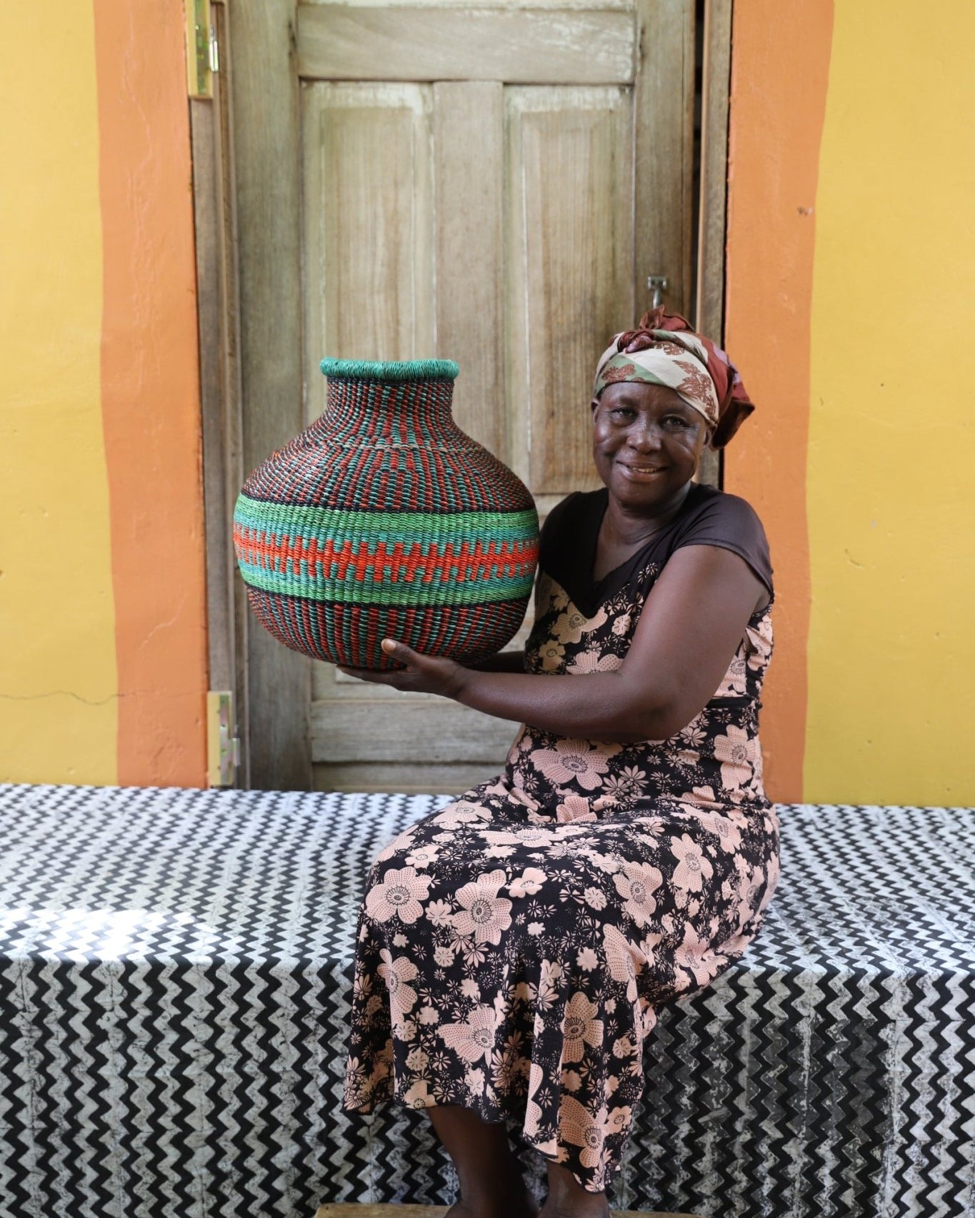 Woman holding a colorful woven basket in a room with yellow walls and a patterned tablecloth.