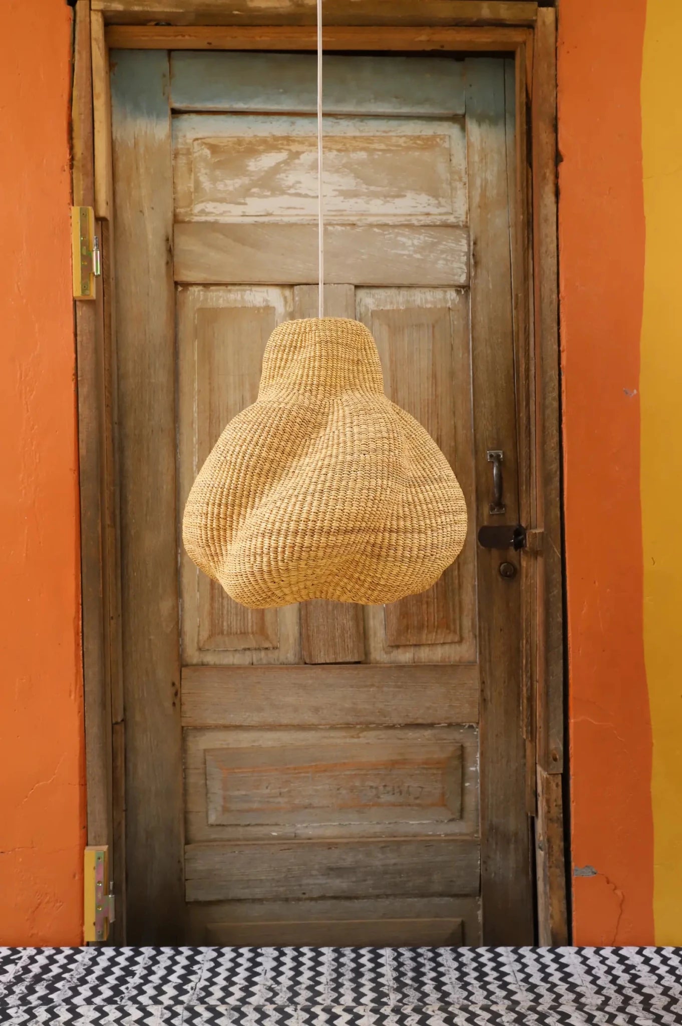 Woven pendant light hanging in front of a wooden door with an orange wall.