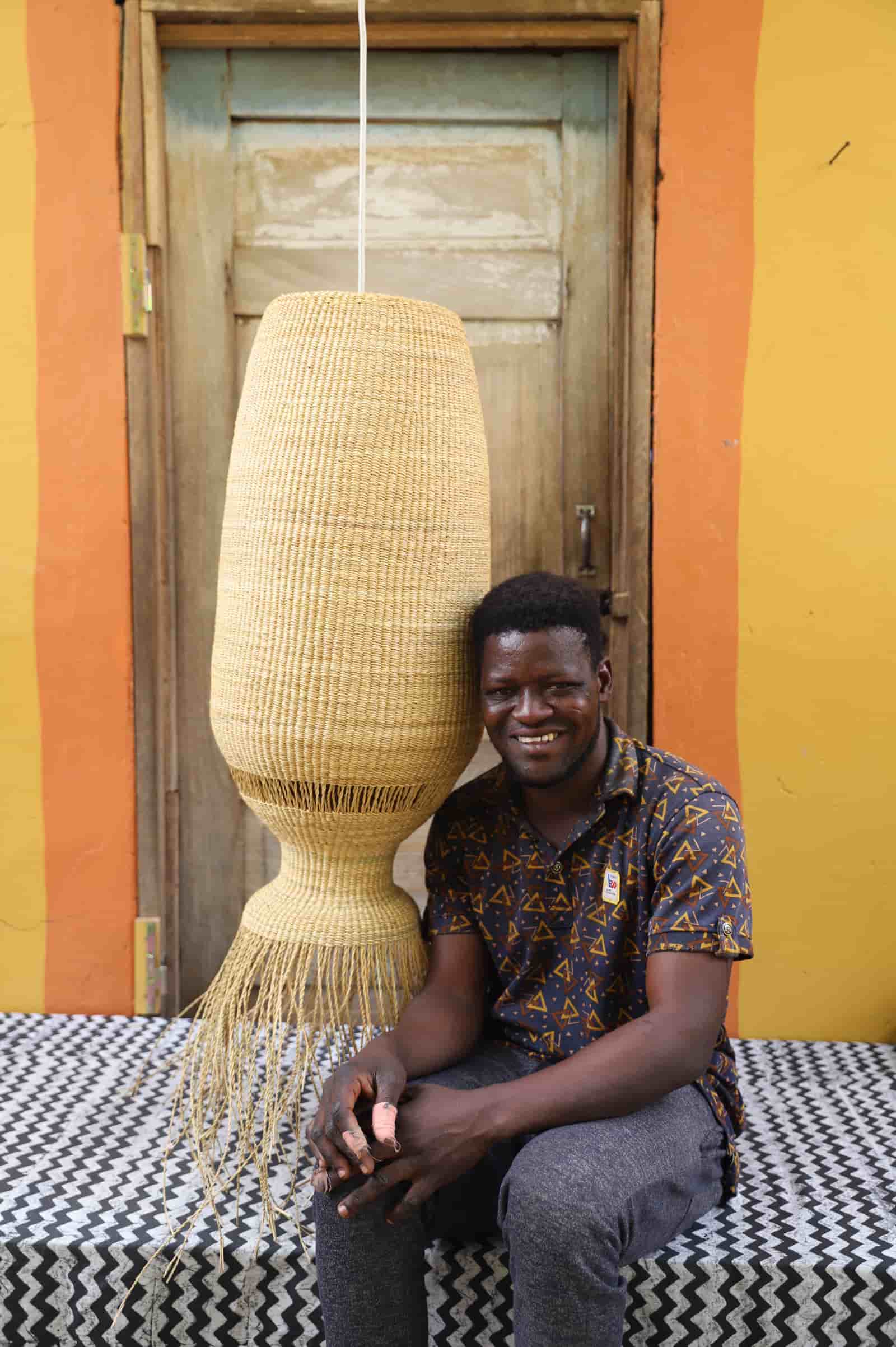 Man sitting next to a large woven lamp against a colorful wall.