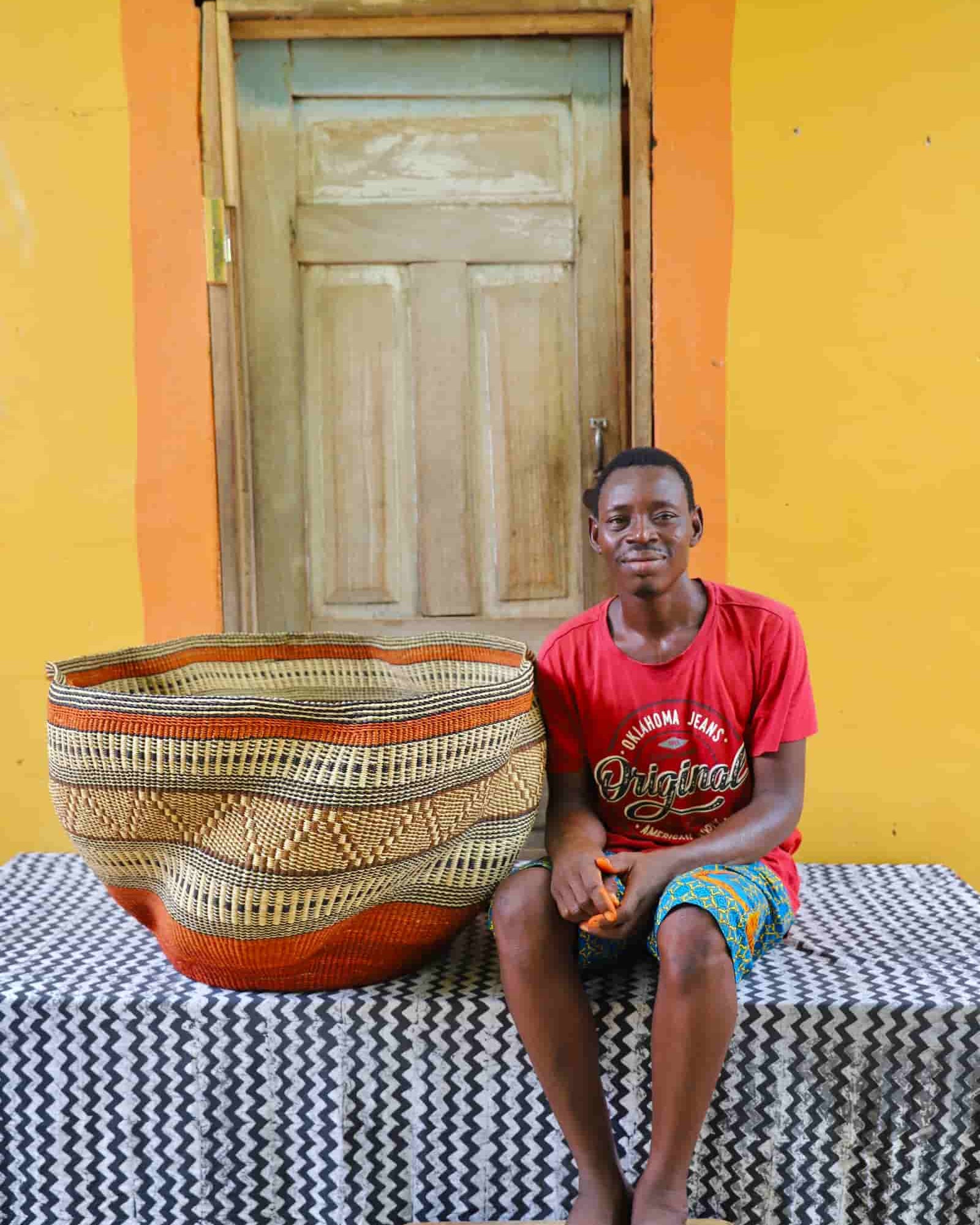 Man sitting on a table with a large woven basket in front of a yellow wall and wooden door.