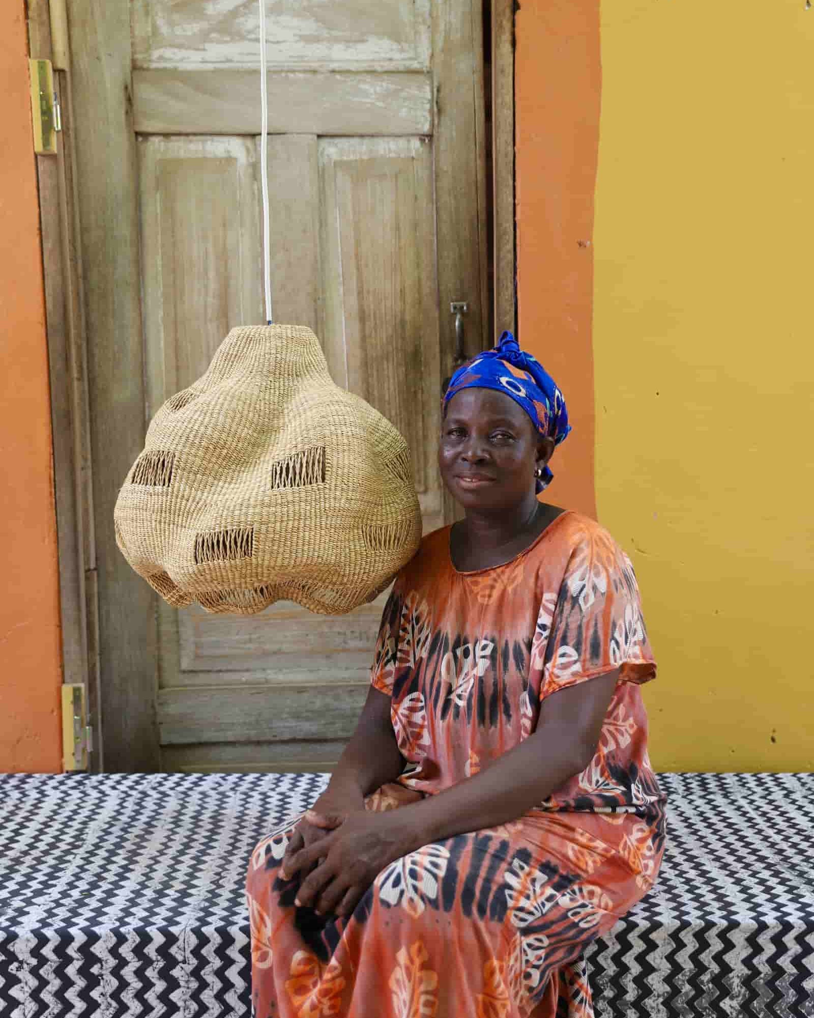 Woman sitting on a bench with a woven basket, wearing an orange dress and blue headscarf, against a colorful wall.