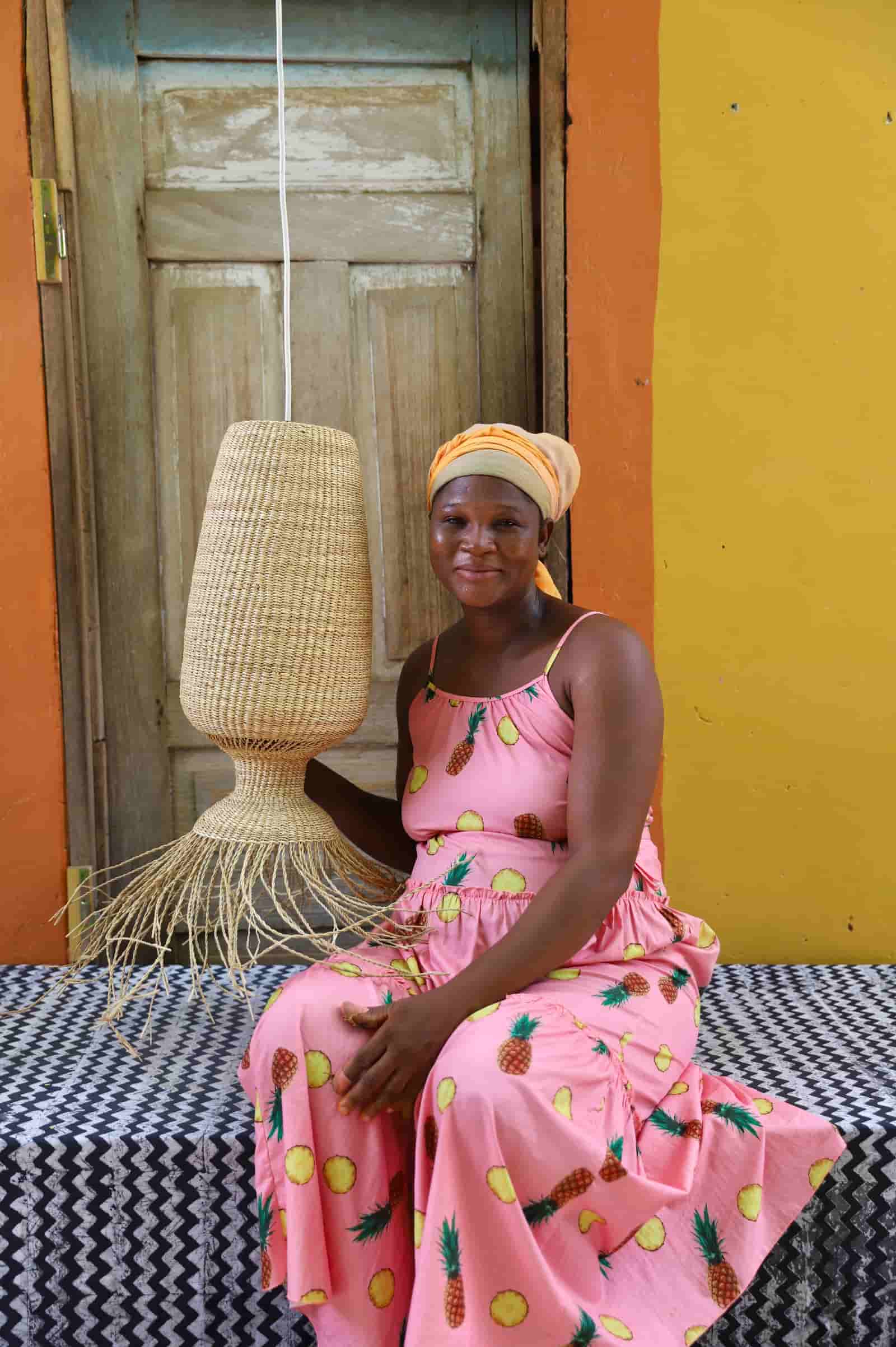 Woman in a pink dress with pineapple pattern holding a woven lampshade against a colorful background.