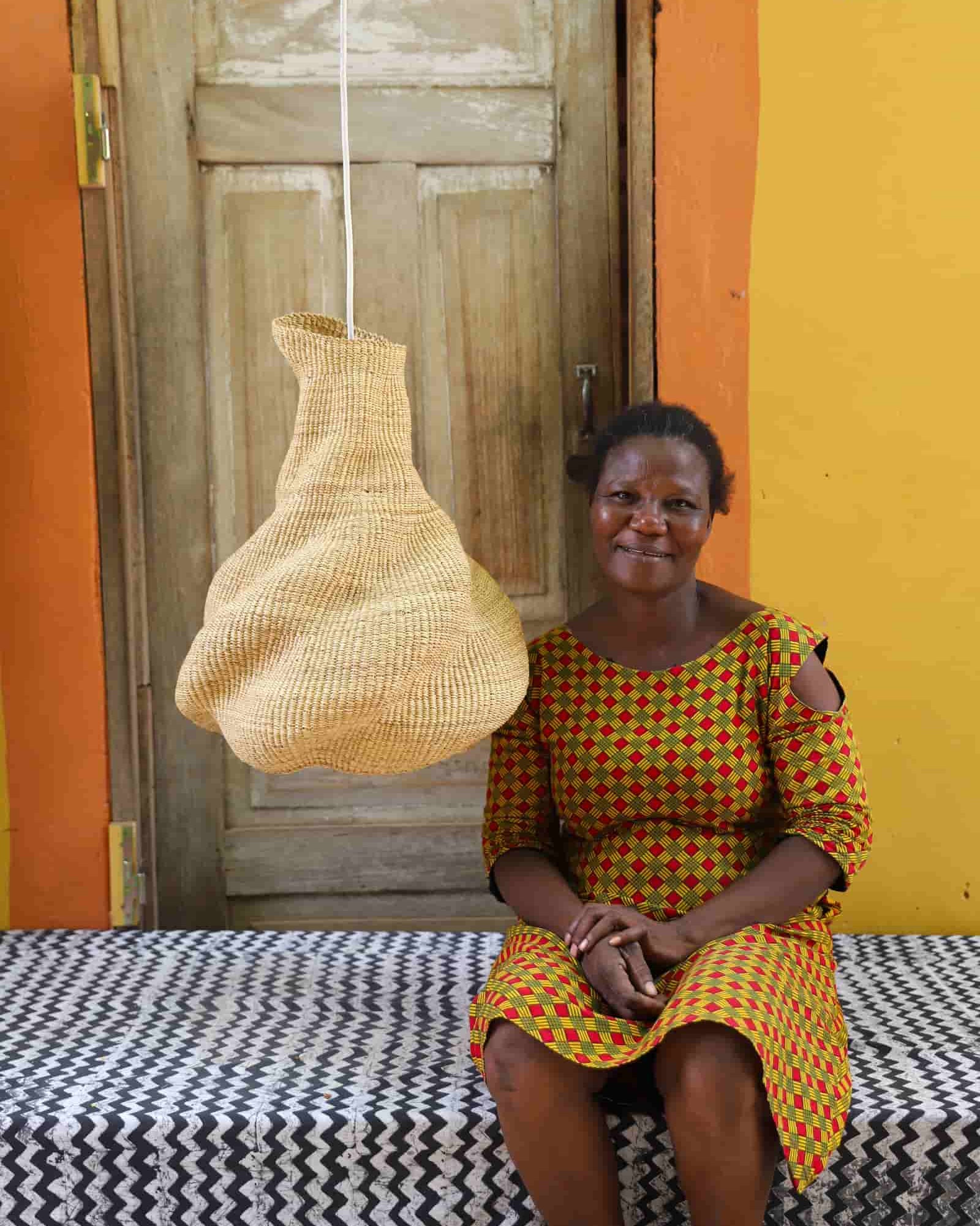 Woman sitting on a bench with a woven basket hanging above her against an orange wall.