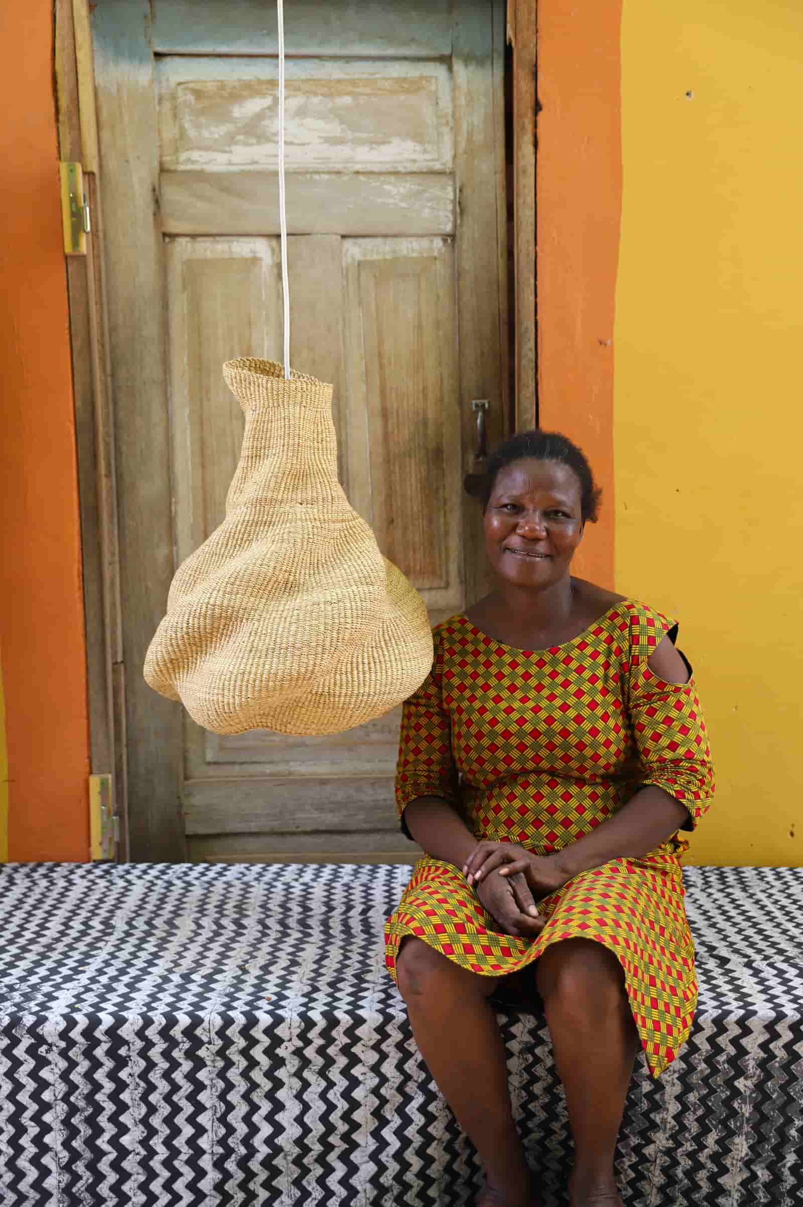 Woman sitting on a bench with a woven basket hanging above her against an orange wall.