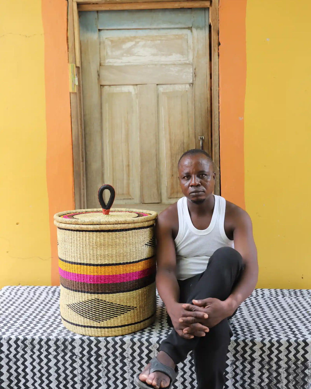 Handwoven Bolga laundry basket with pink and orange bands, beside a seated man in a white vest.
