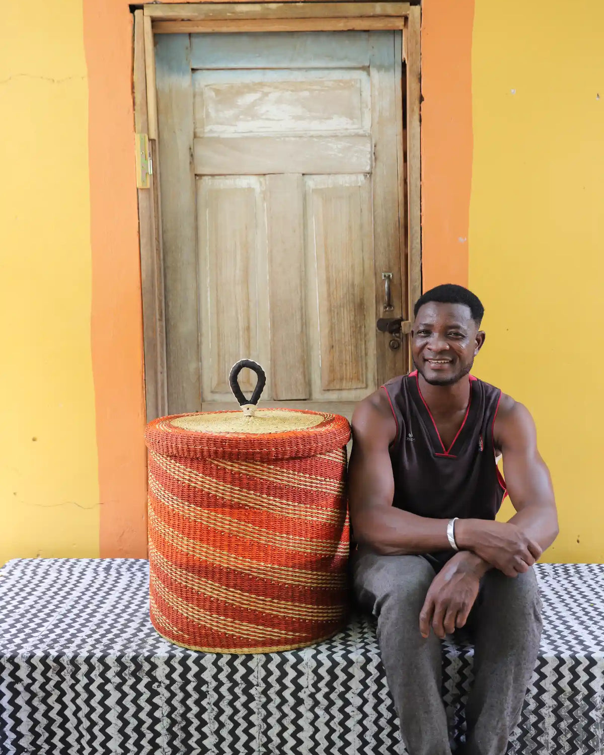 Handwoven Bolga laundry basket dyed rust-orange, beside a seated man in a dark vest.