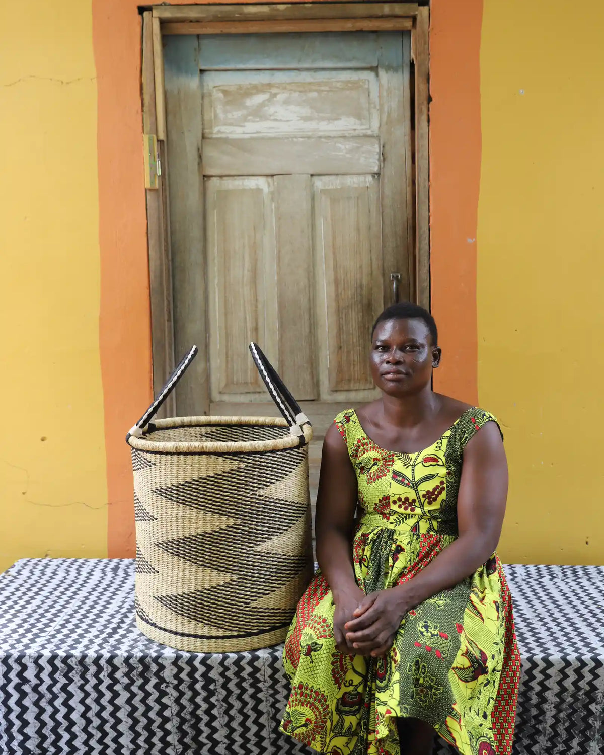 Medium handwoven Bolga laundry basket in natural with black sawtooth stripes, beside a seated woman in green.
