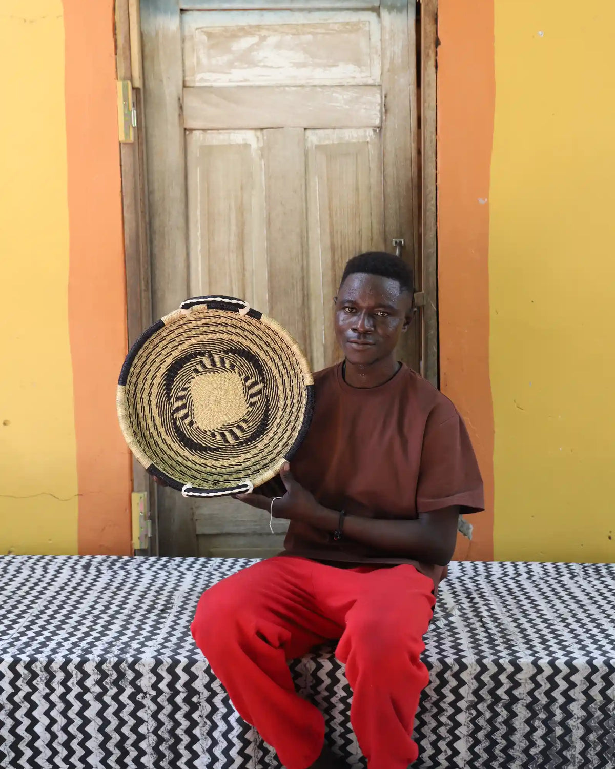 Handwoven Bolga wall tray disc in natural with yellow centre, held by a seated man in red.