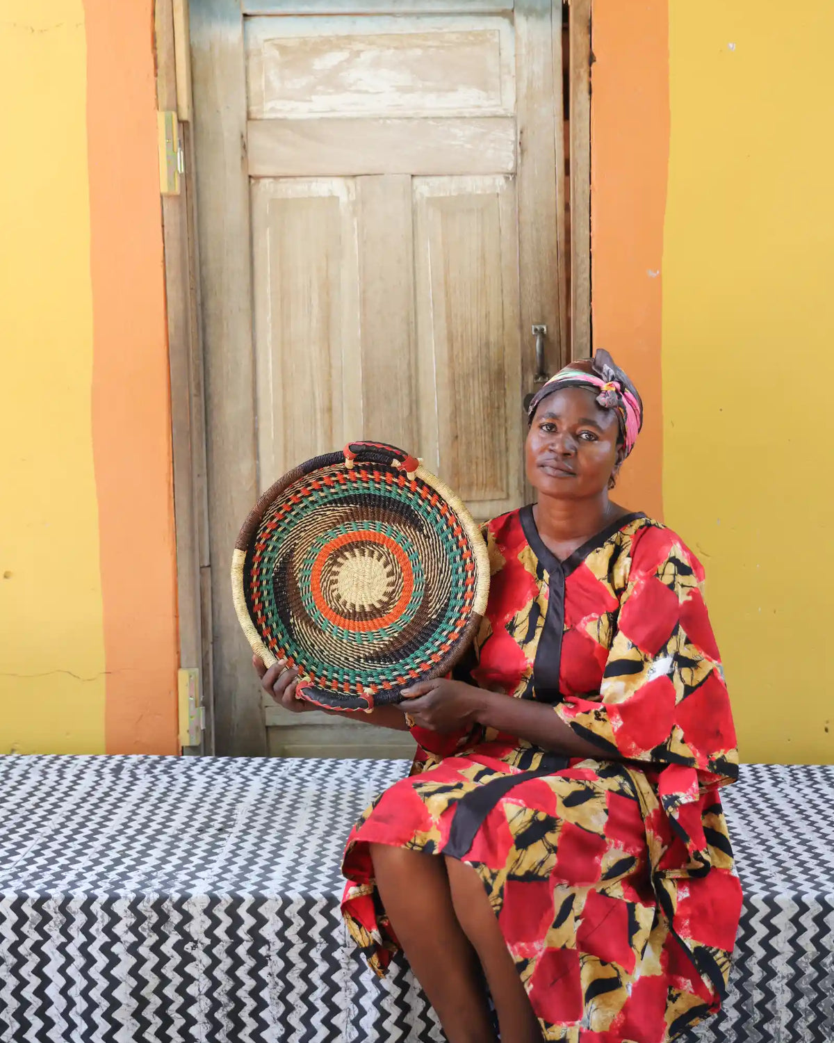 Handwoven Bolga wall tray disc with green and orange bands, held by a seated woman in red floral dress.