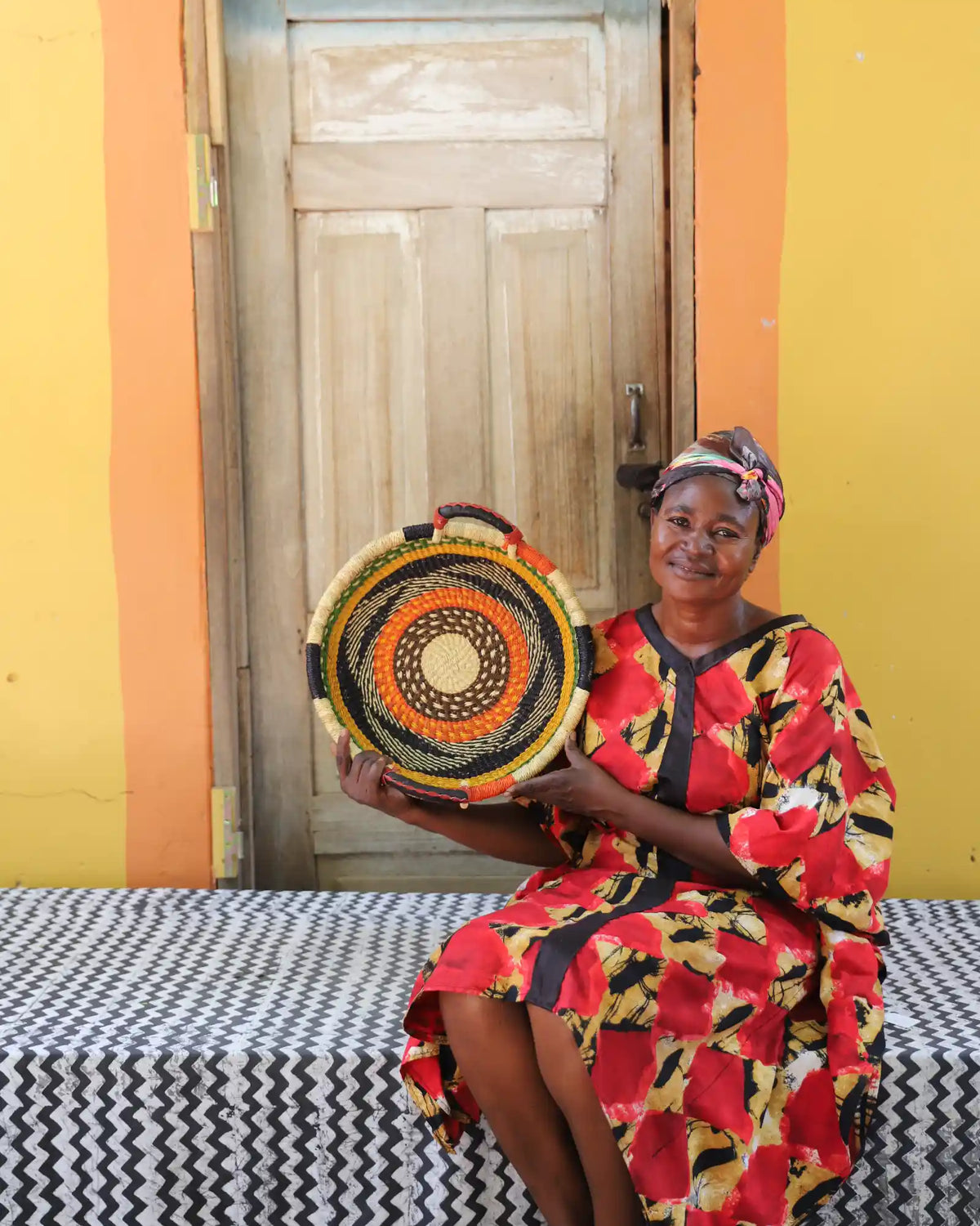 Handwoven Bolga wall tray disc with orange and black bands, held by a seated woman in red floral.