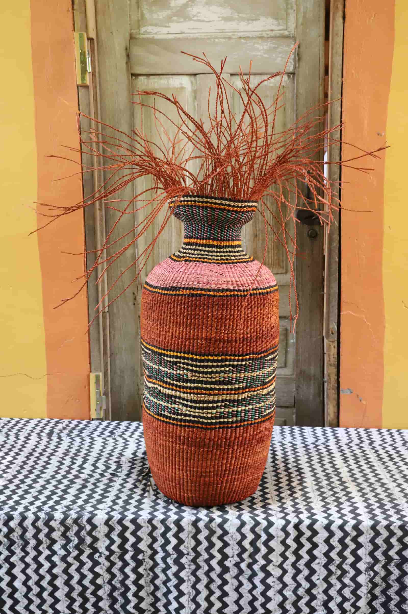 Decorative woven vase with red branches on a tablecloth against a colorful wall.