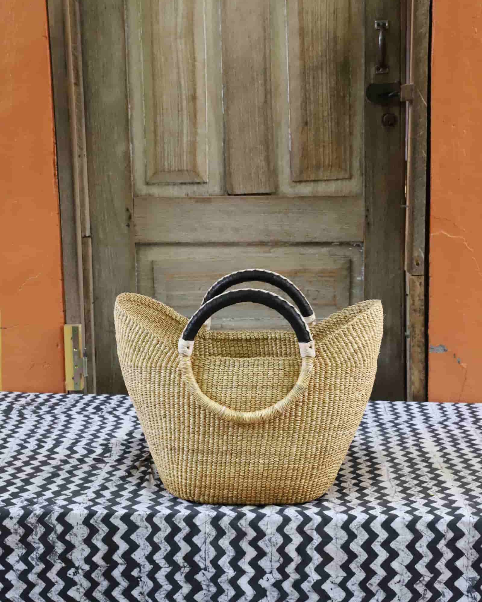 Woven handbag with black handles on a tablecloth in front of a wooden door.