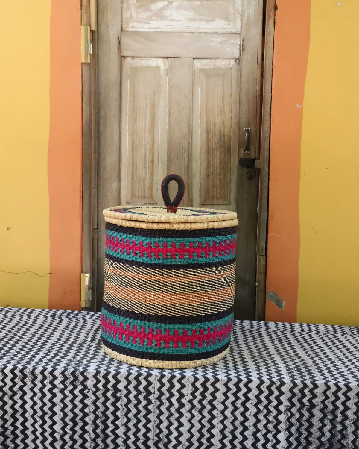 Handwoven Bolga laundry basket with coiled rim and lid in natural straw with fine pink, black and orange bands.