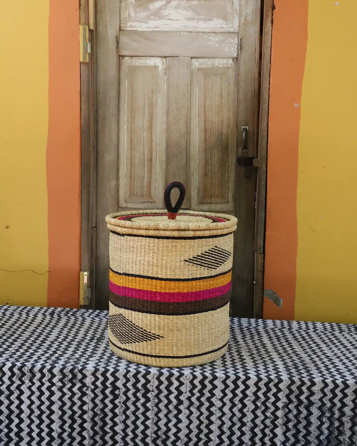 Handwoven Bolga laundry basket with coiled rim and lid in natural straw with pink and orange bands and dark diamond.