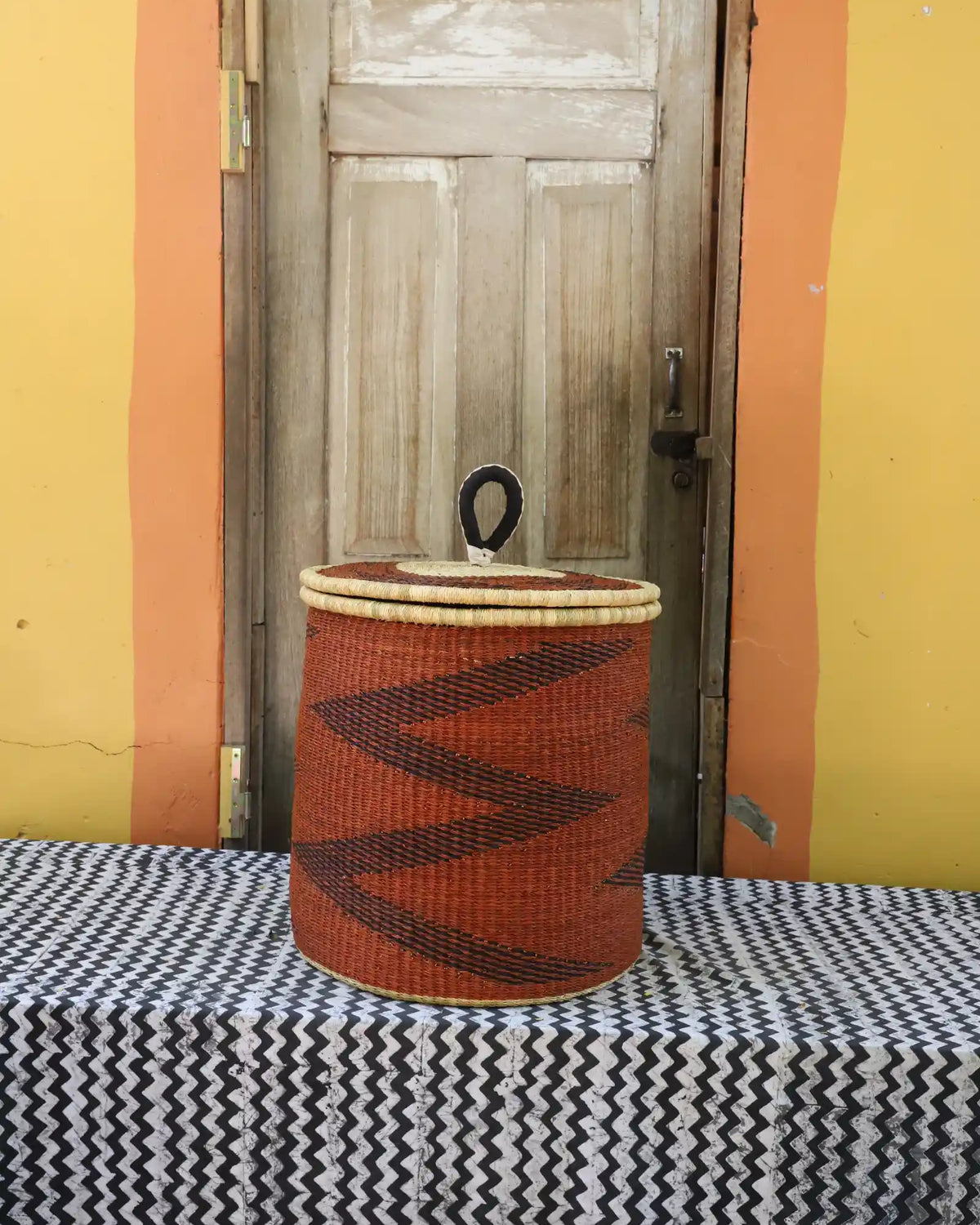 Handwoven Bolga laundry basket with coiled rim and lid dyed rust-orange with a dark zigzag pattern.