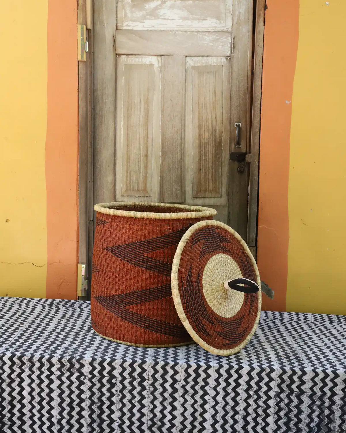 Handwoven Bolga laundry basket with lid removed, rust-orange body with dark zigzag and natural rim lid.