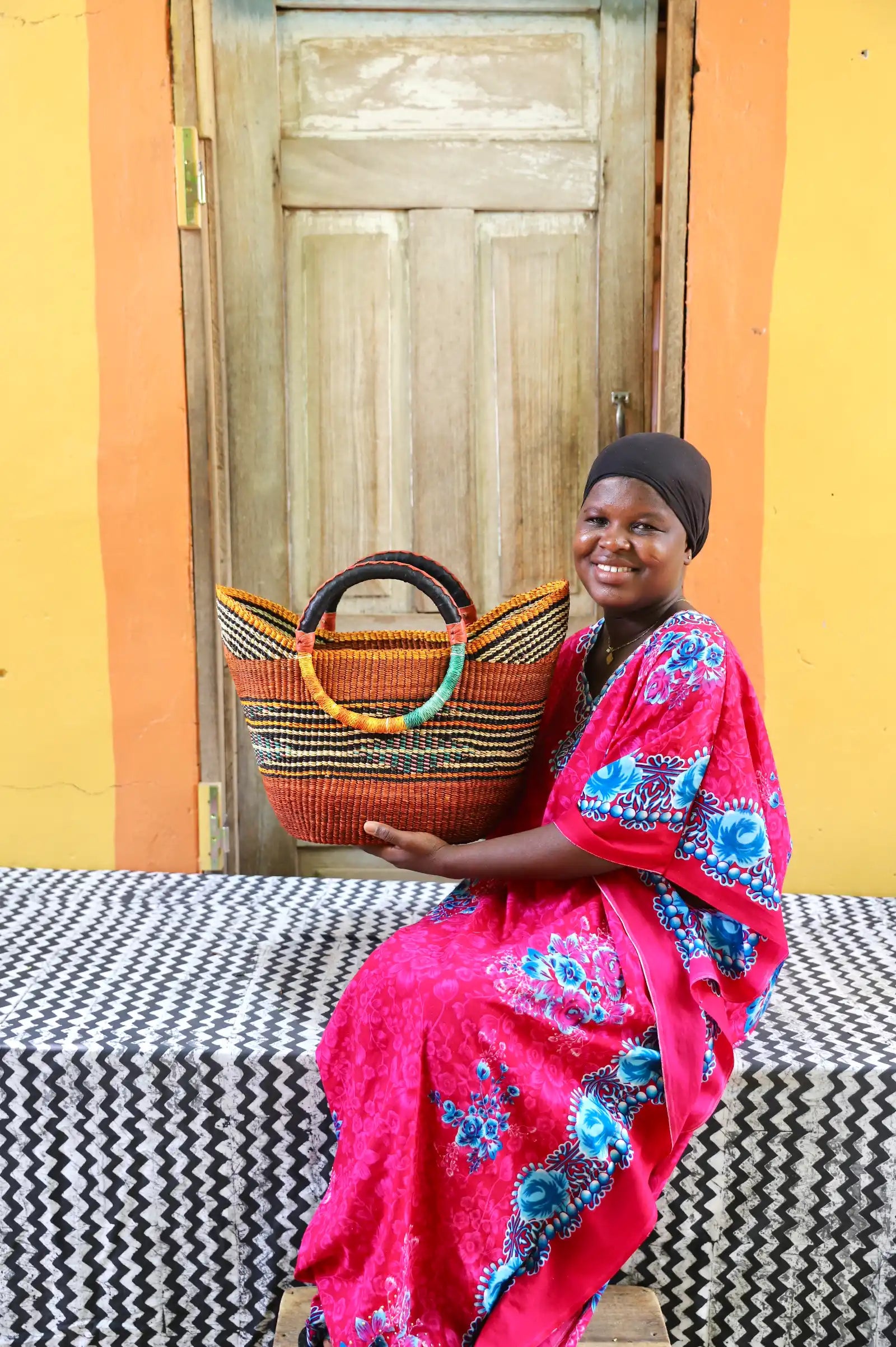 Woman in a bright pink dress with blue patterns holding a woven basket against a colorful wall.