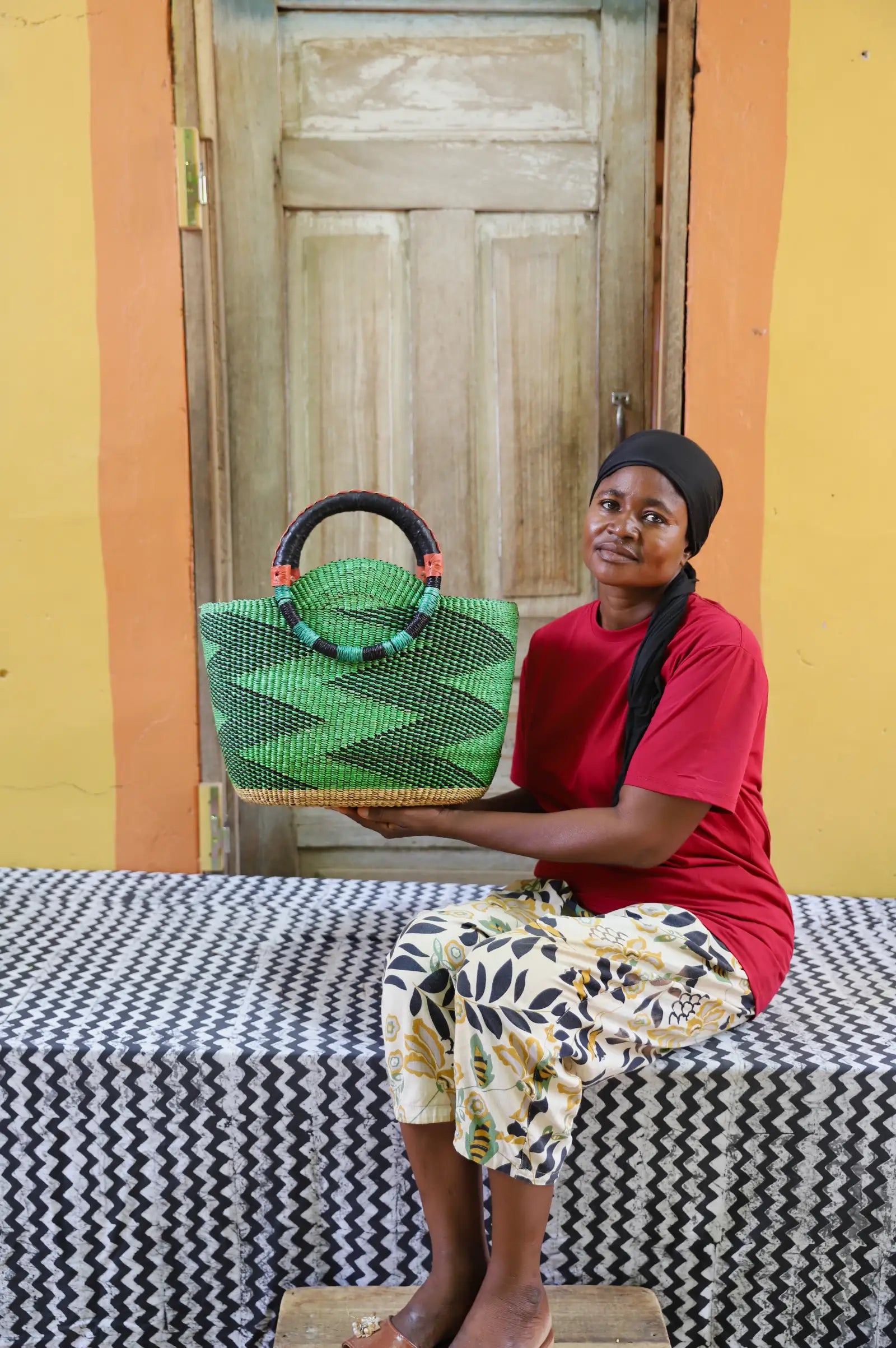Woman holding a green woven handbag in front of a wooden door.