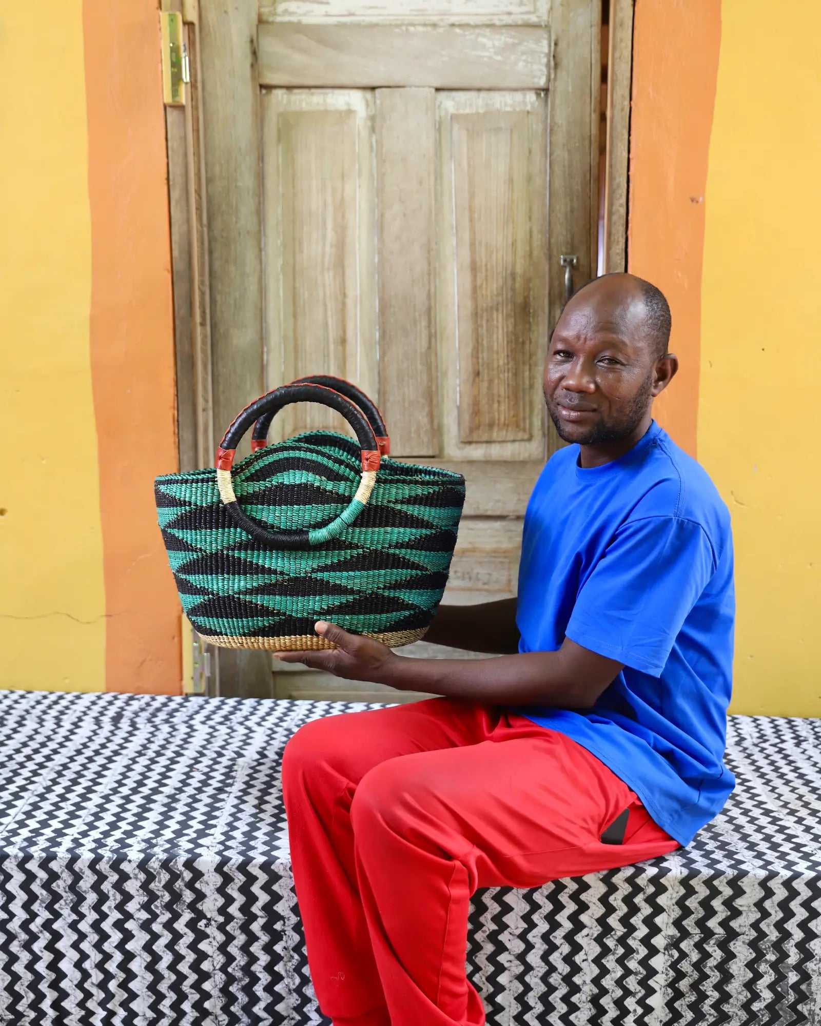 Man holding a woven bag in front of a wooden door with a colorful wall.