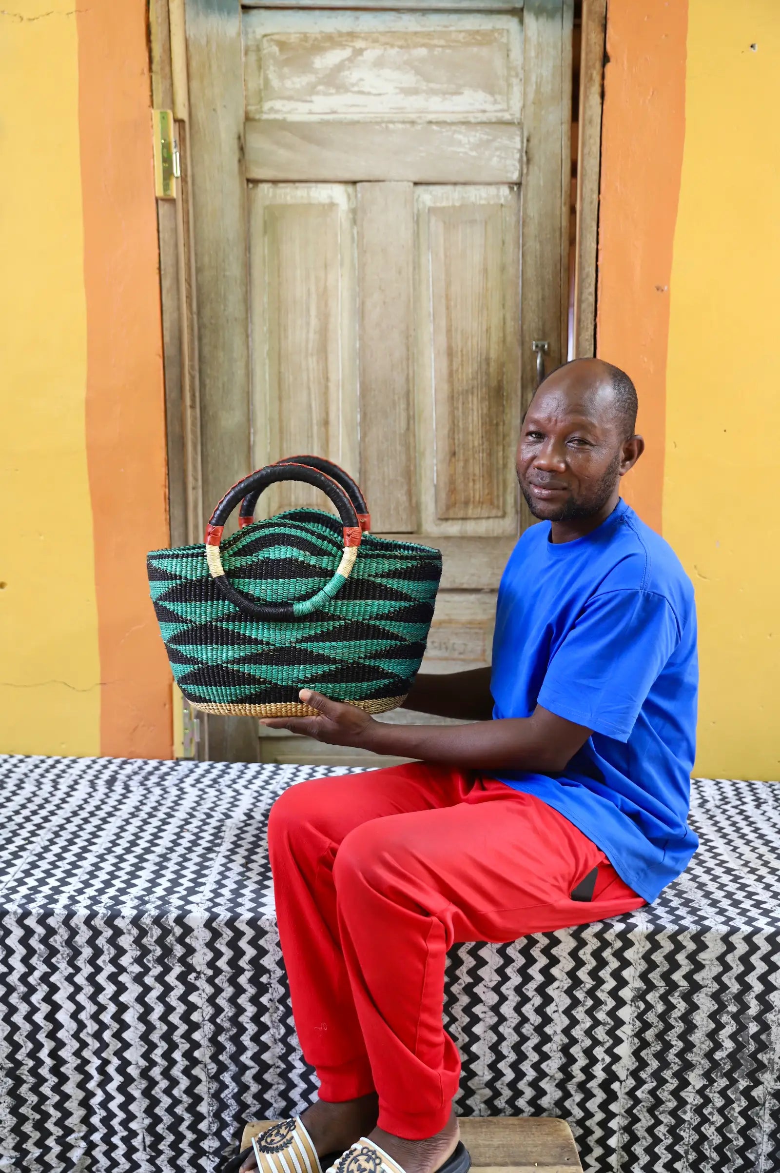 Man holding a woven bag in front of a wooden door with a colorful wall.