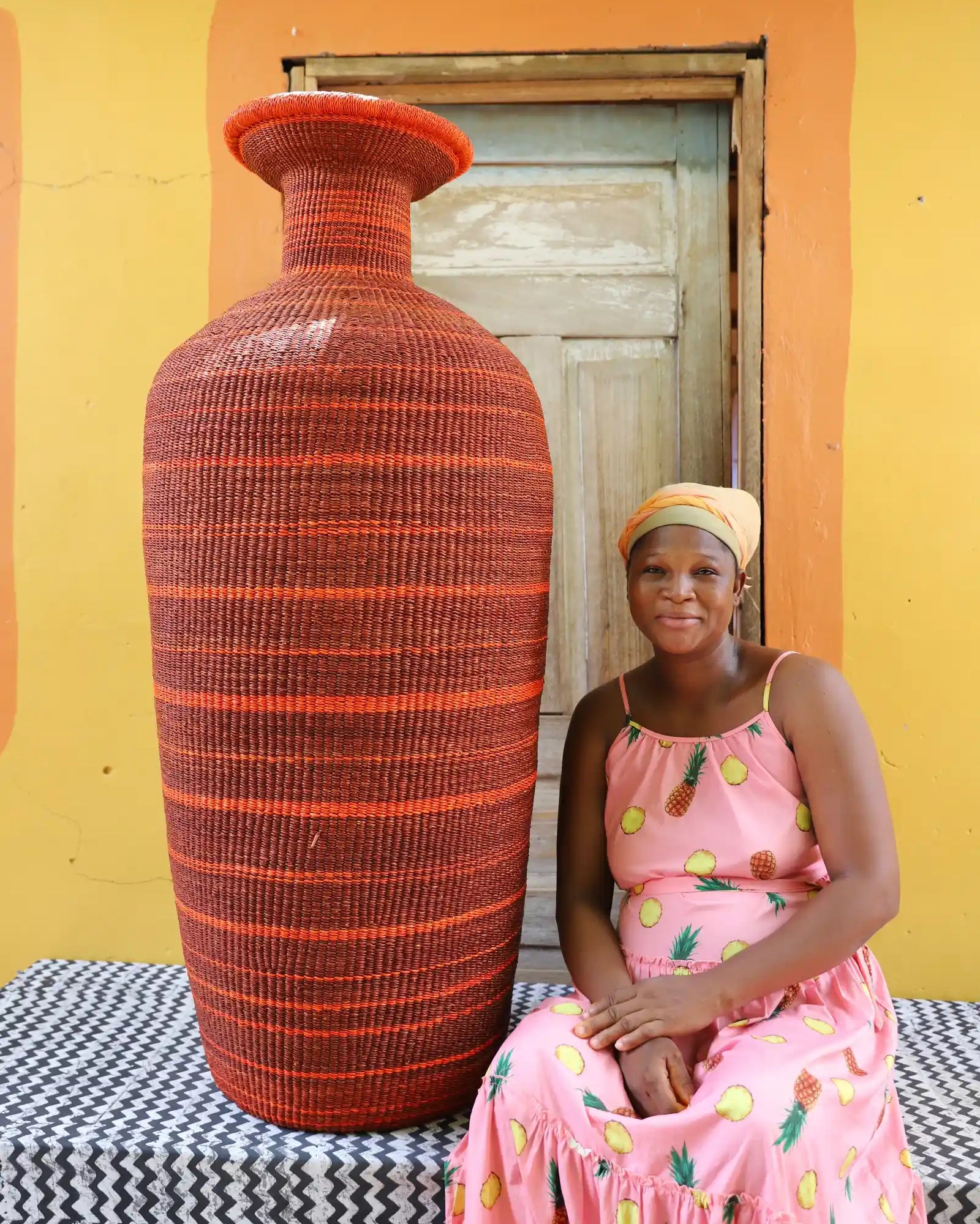 Woman in a pink dress sitting next to a large red woven vase against a yellow wall.