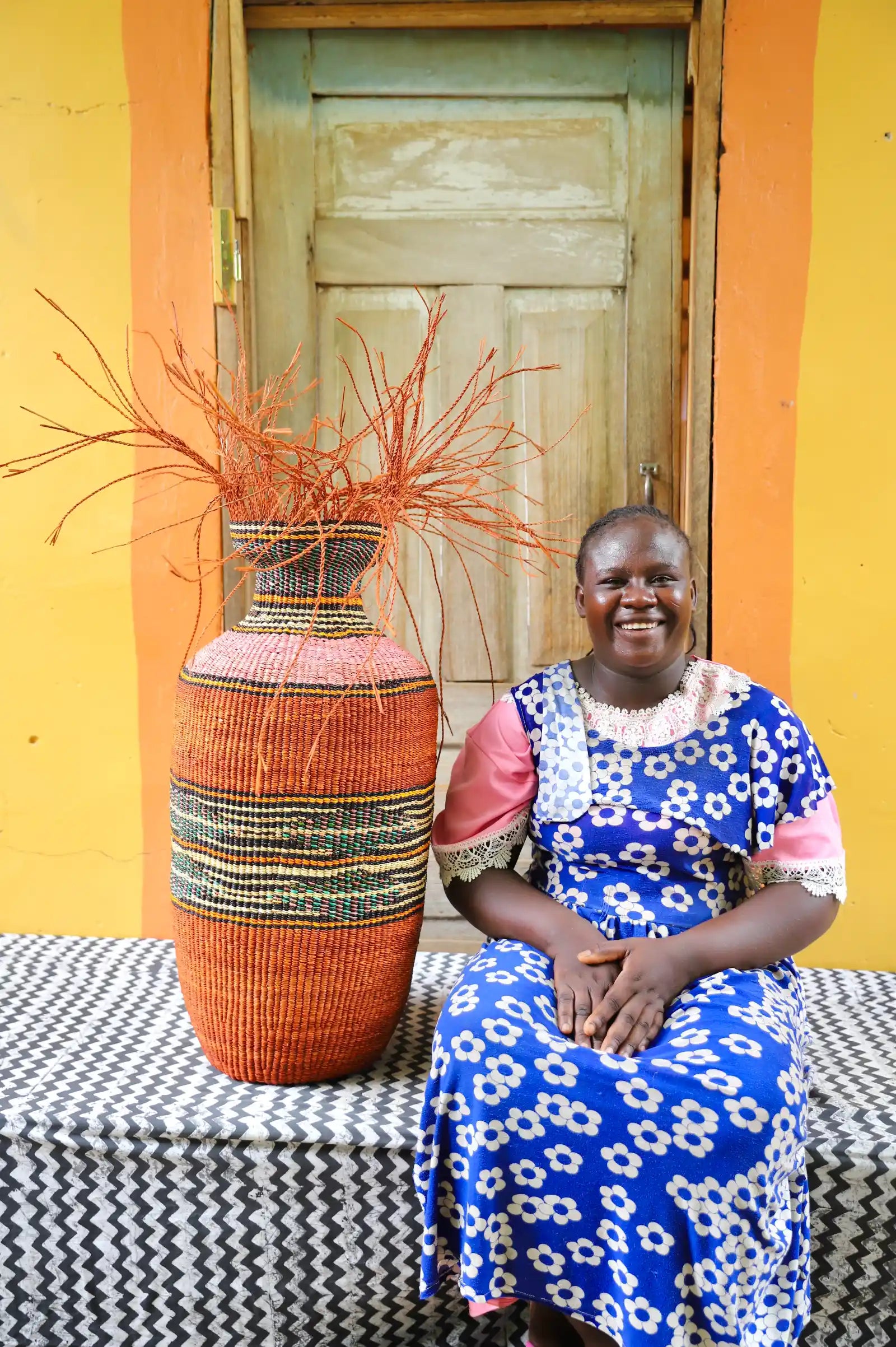 Woman sitting next to a large woven vase against a colorful wall.